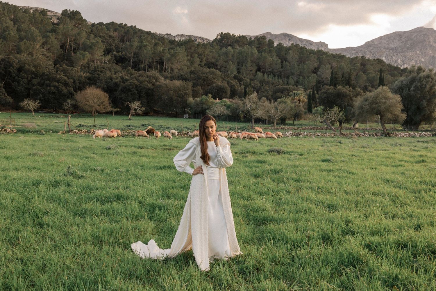 An elegant white gown flows in the breeze against a mountainous countryside backdrop with sheep.