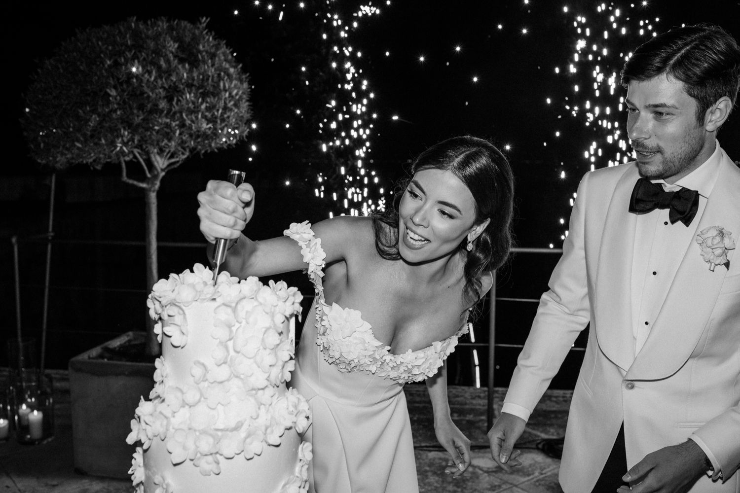 Black and white photo of bride and groom cutting wedding cake with sparklers in background.