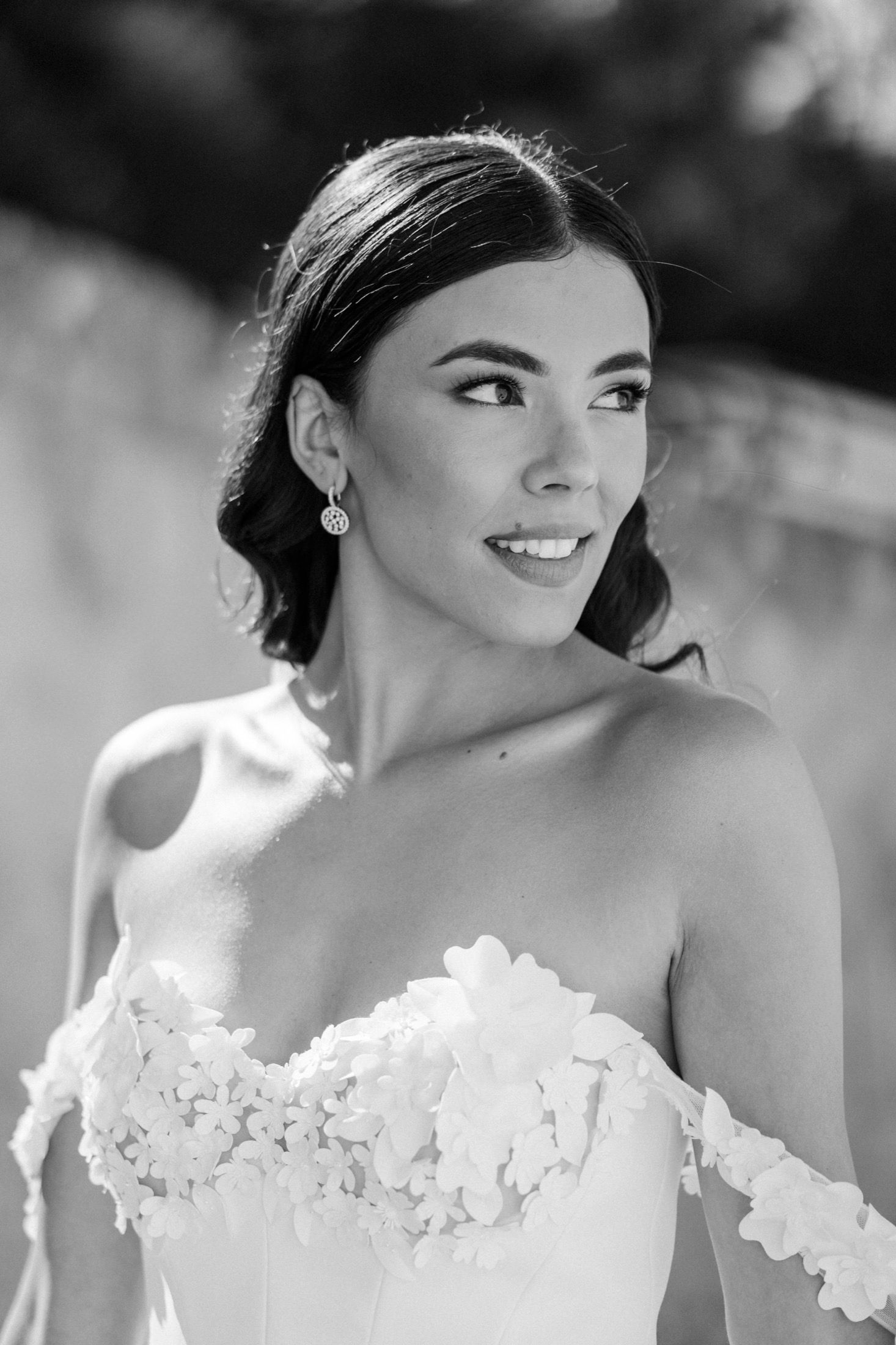 Black and white portrait of a smiling bride in a floral off-shoulder wedding gown with elegant drop earrings.