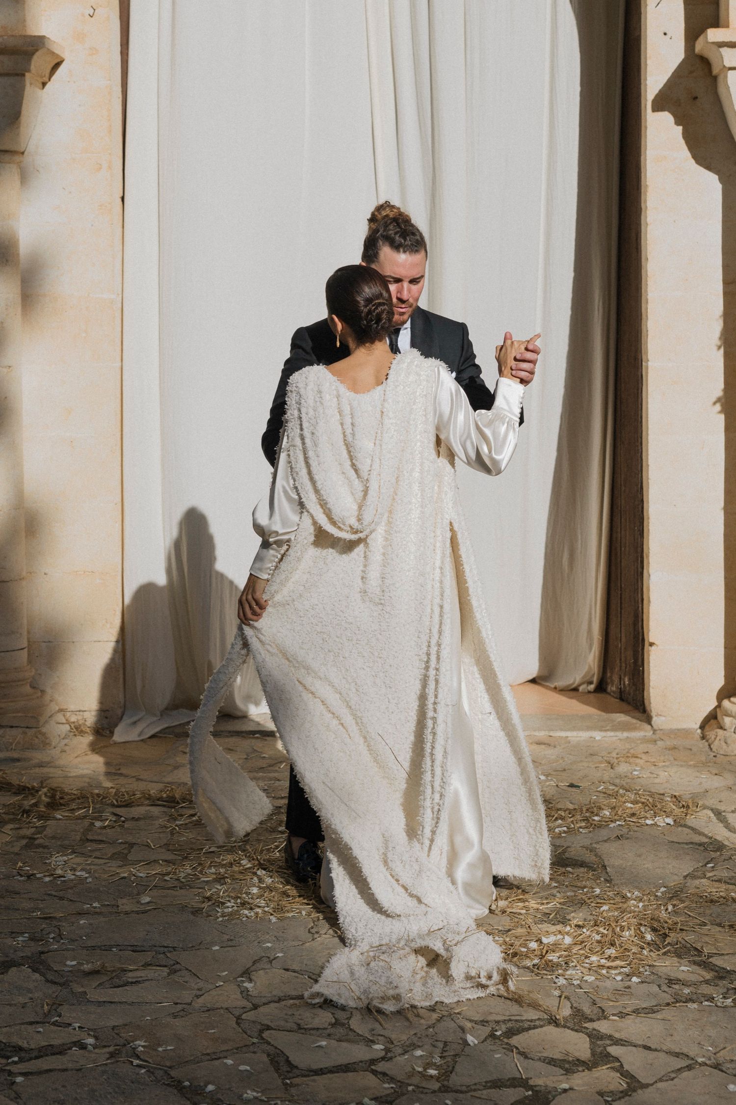 Wedding couple dancing playfully through stone colonnade with flowing white dress creating elegant motion.