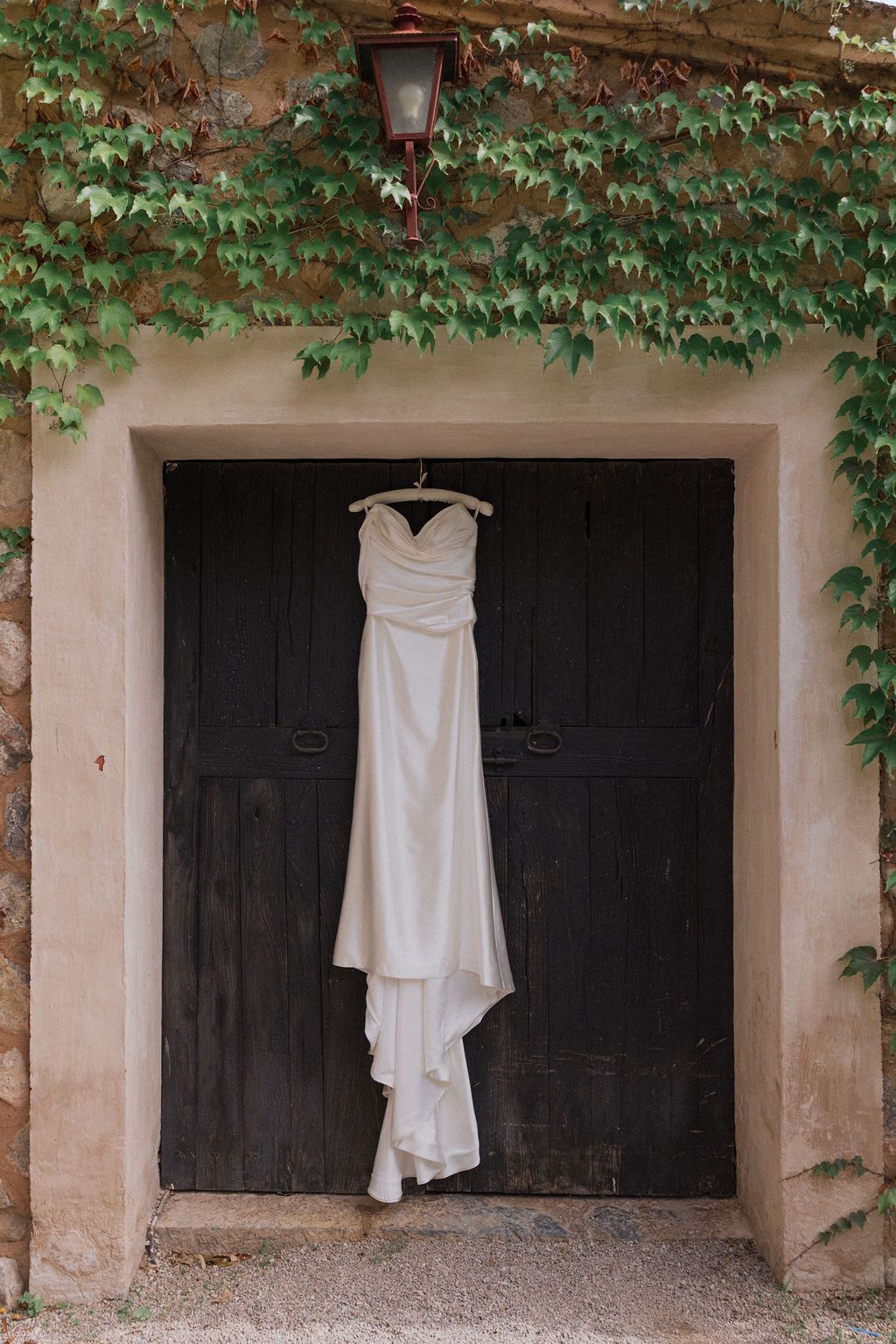 White wedding dress hanging against dark wooden doors with ivy growing above.