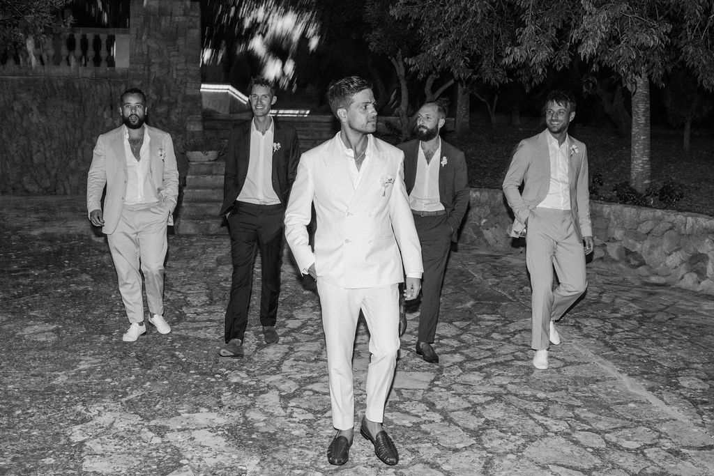 Groomsmen in light colored suits celebrate on cobblestone ground at night.