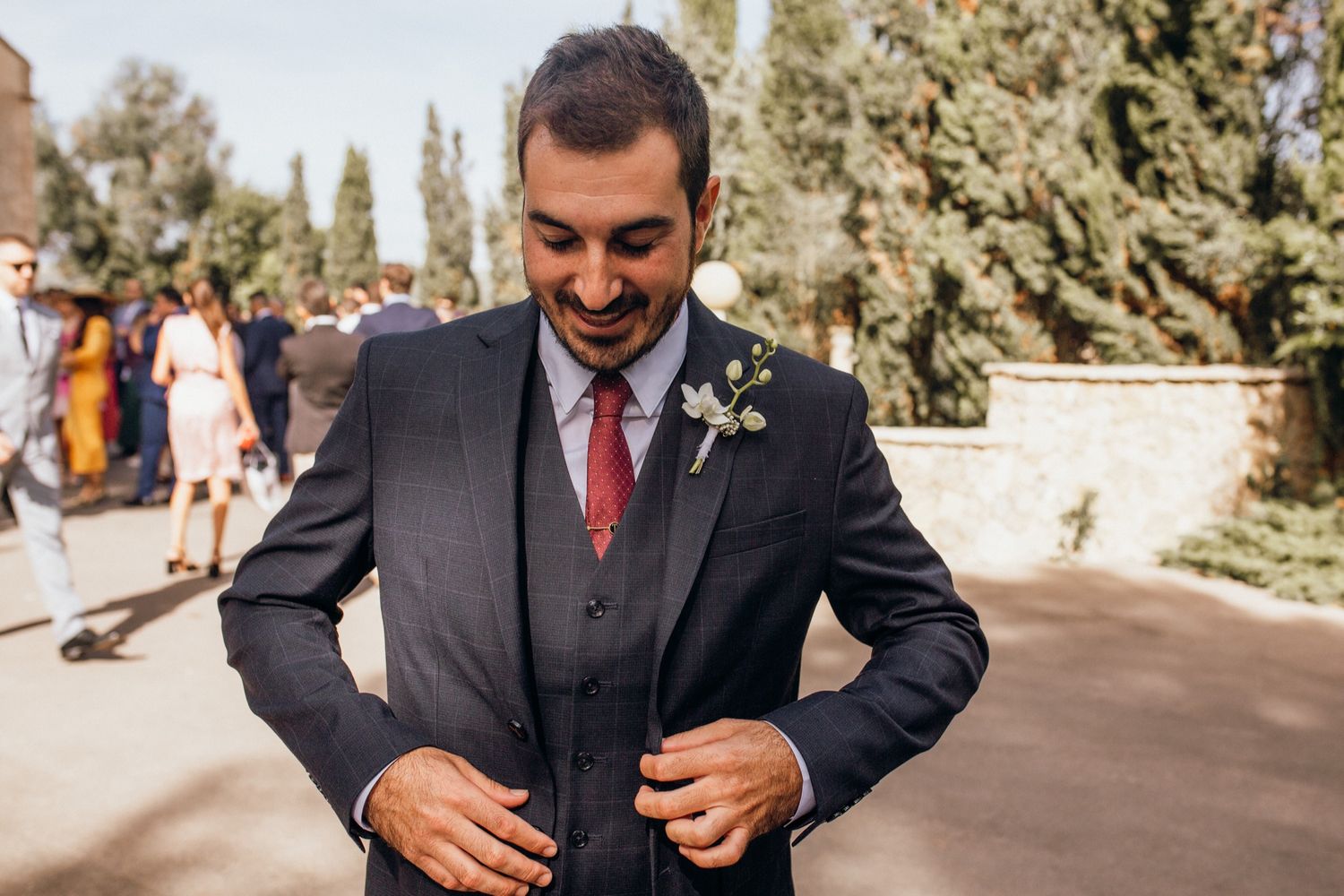 Groom in dark suit adjusts his tie while standing outdoors with trees in the background.