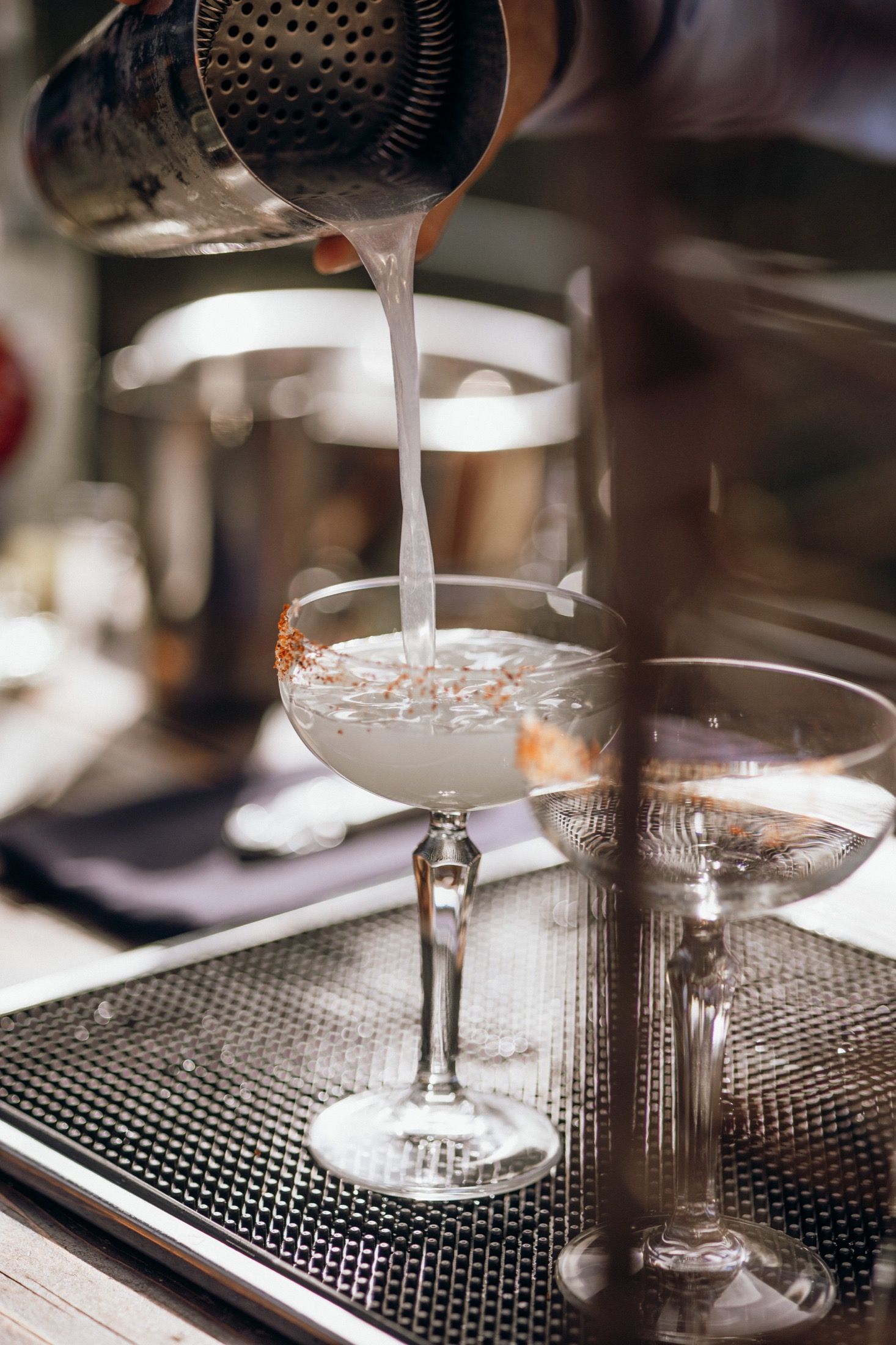 Sequence of cocktail glasses being filled with white liquid on a metal bar mat surface.