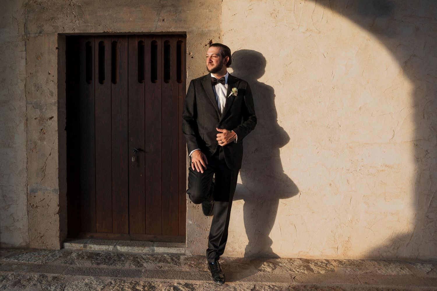 A groom in a classic black tuxedo poses against a textured wall with dramatic shadows.