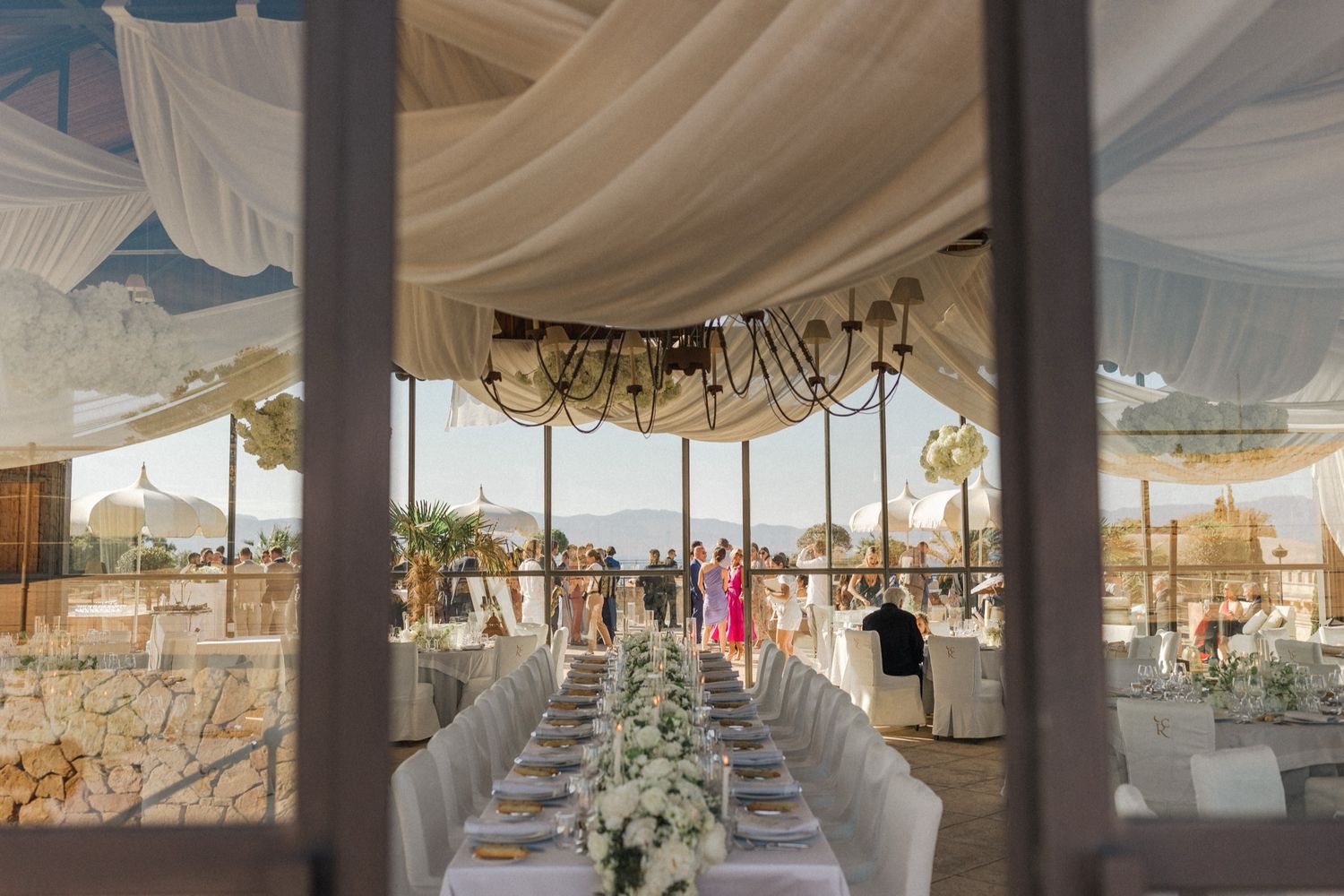 Elegant outdoor wedding reception tent with white draping, long decorated tables, floral centerpieces, and ocean views.