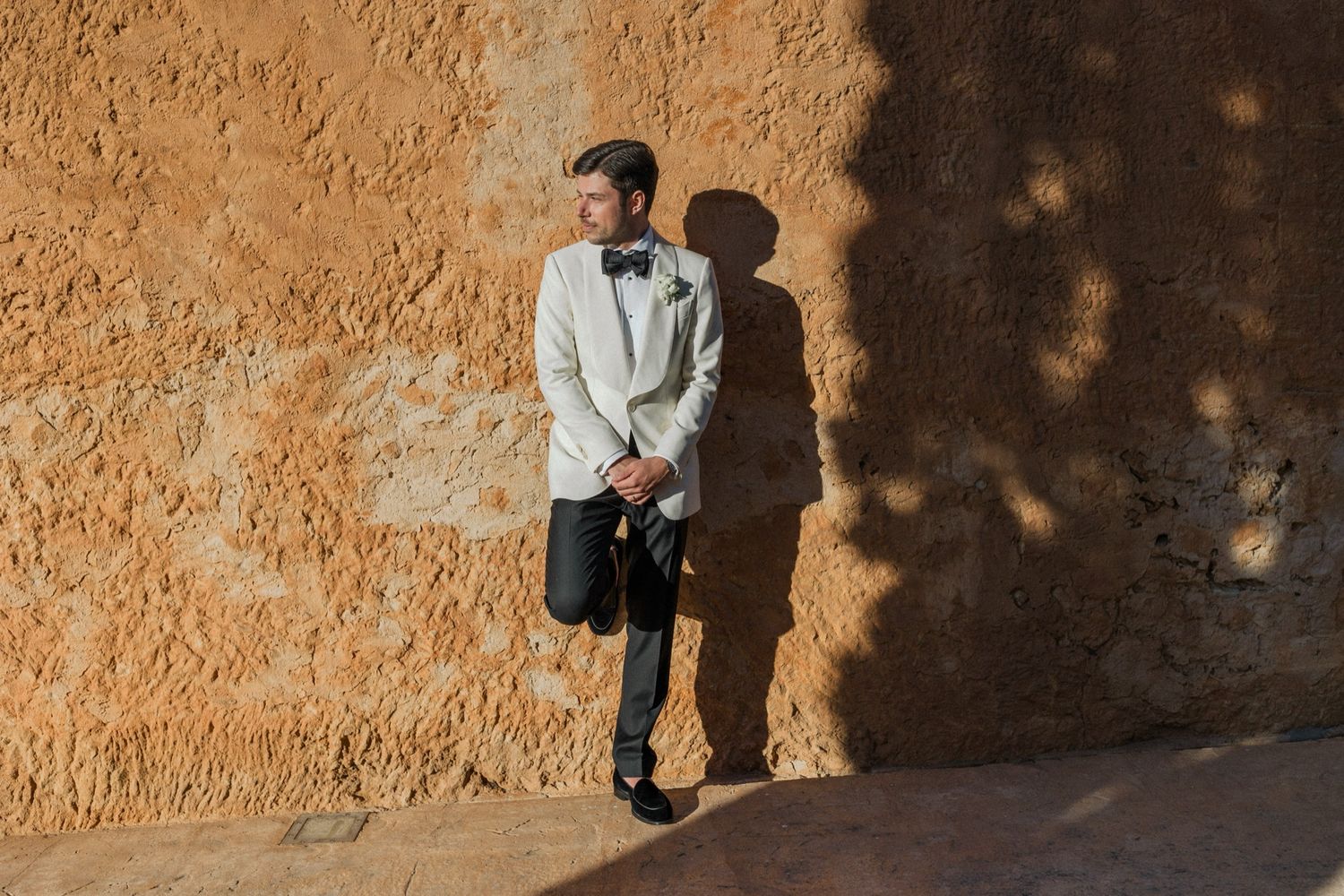 Man in white tuxedo jacket leaning against rustic orange stone wall with dramatic shadows.