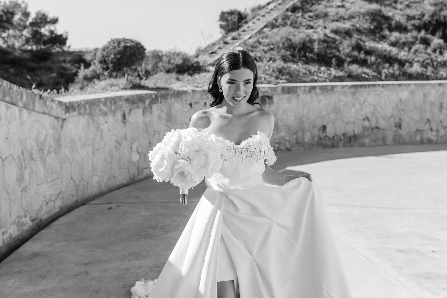Black and white photo of a smiling bride in an off-shoulder gown holding a white bouquet outdoors.
