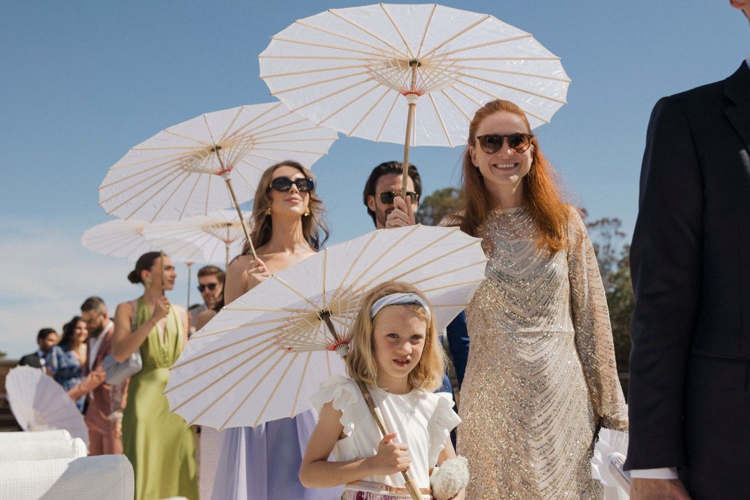 Wedding guests holding white parasols walk in a sunny outdoor ceremony, featuring a flower girl and woman in a sequined dress.