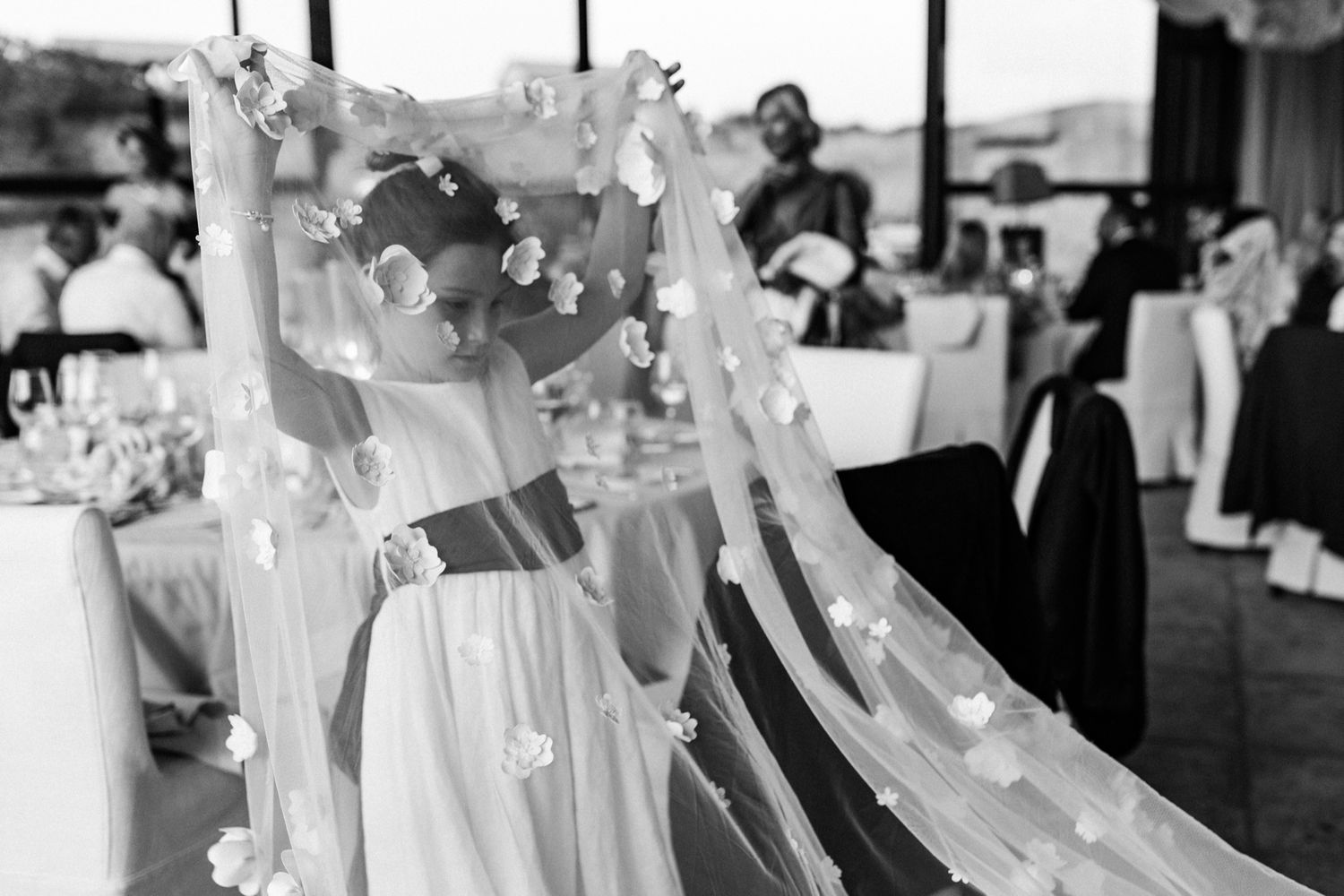 Black and white photo of a flower girl in a floral veil at a wedding reception venue.