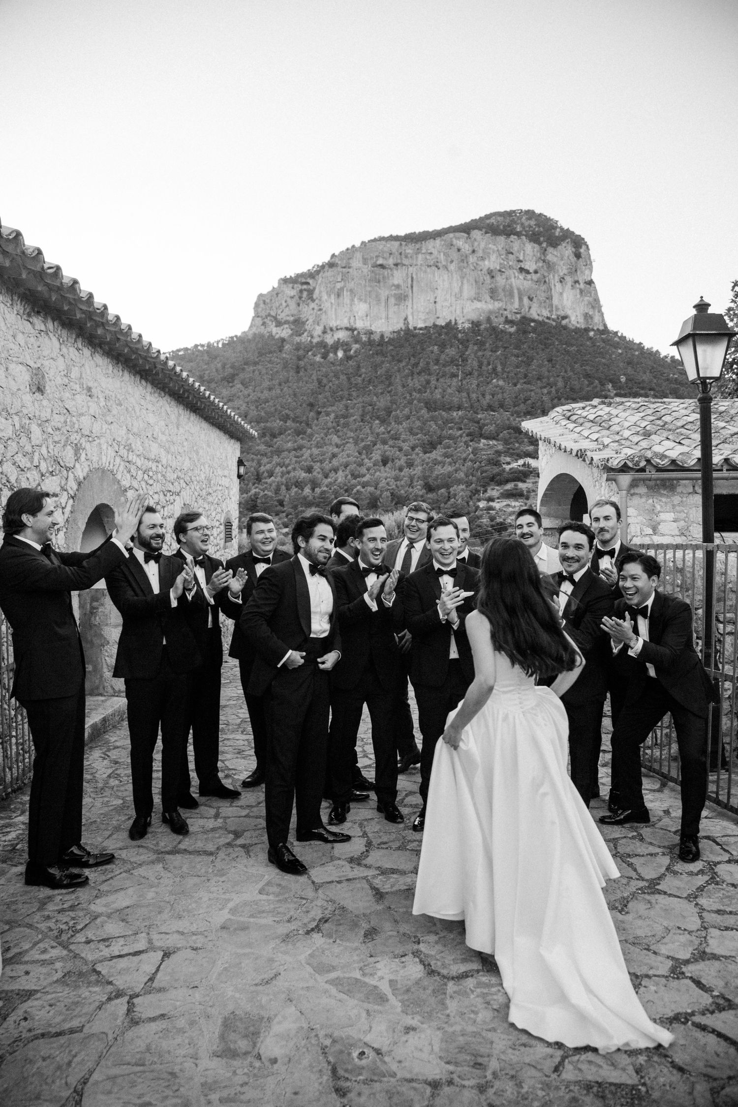 A wedding celebration takes place on a stone terrace with a dramatic mountain backdrop in black and white.