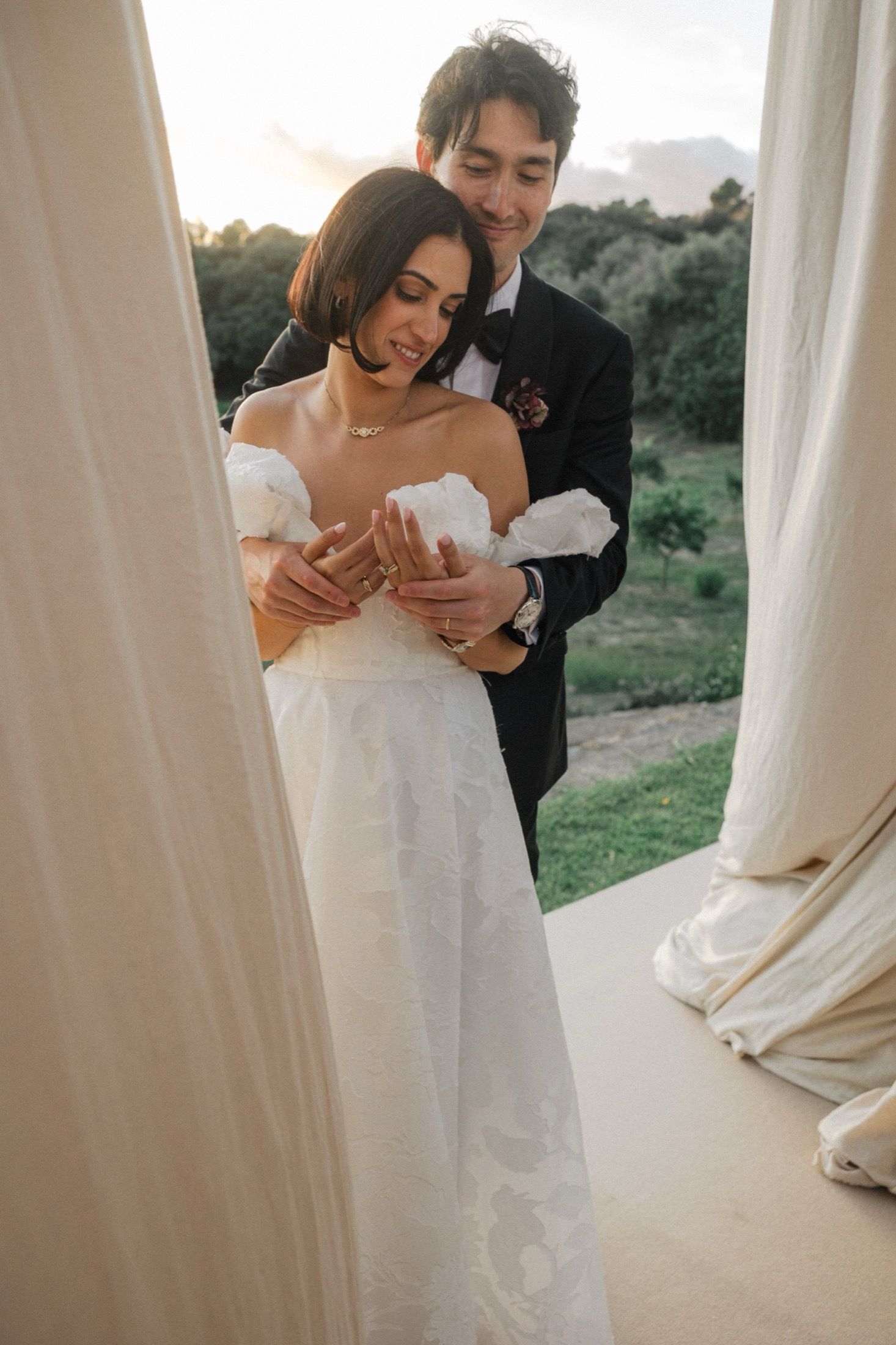 A couple shares an intimate moment in wedding attire on an outdoor terrace with flowing white curtains at sunset.