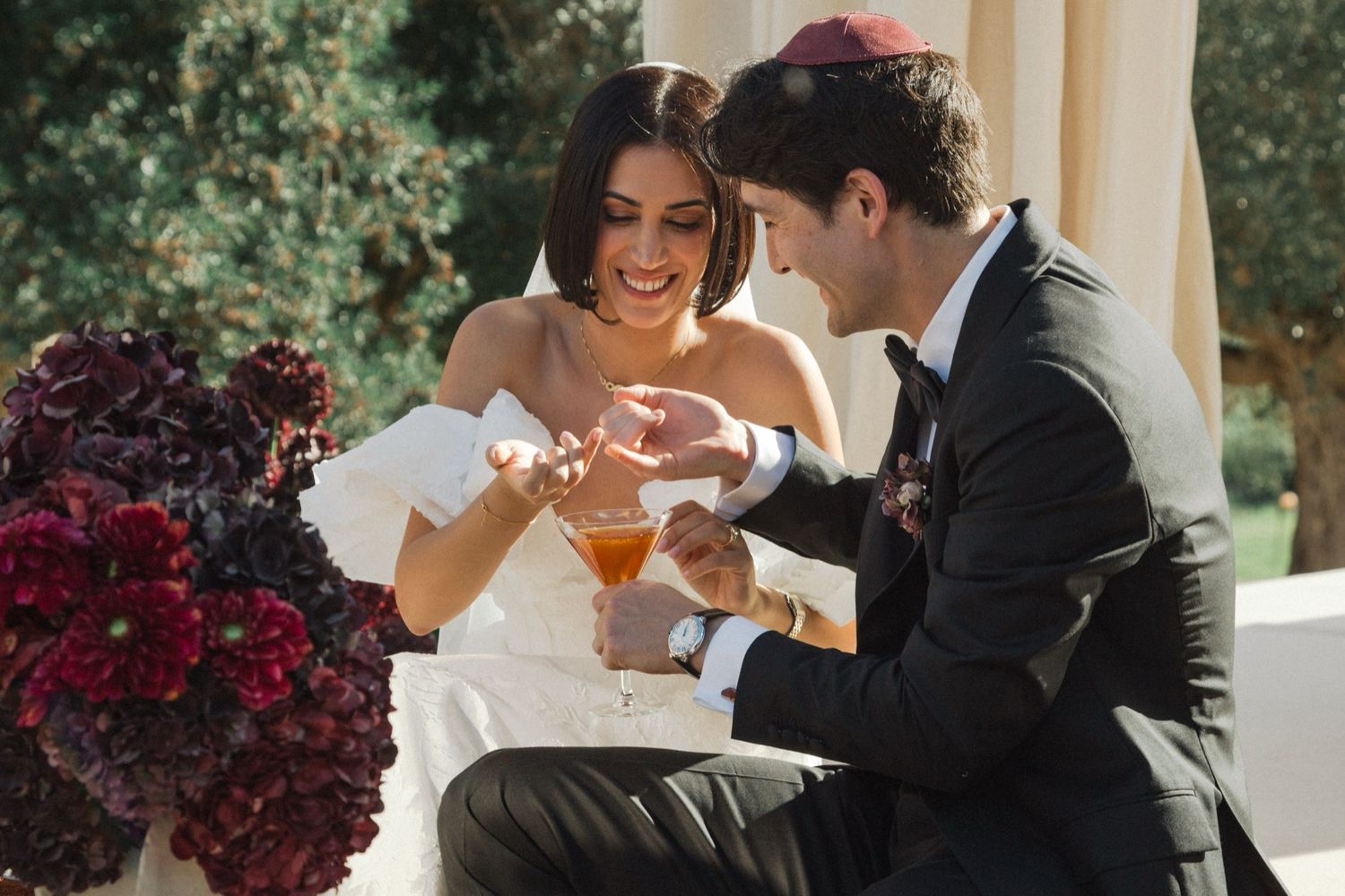 Newlyweds sharing cocktails and smiling together at an outdoor wedding reception with burgundy floral decor.