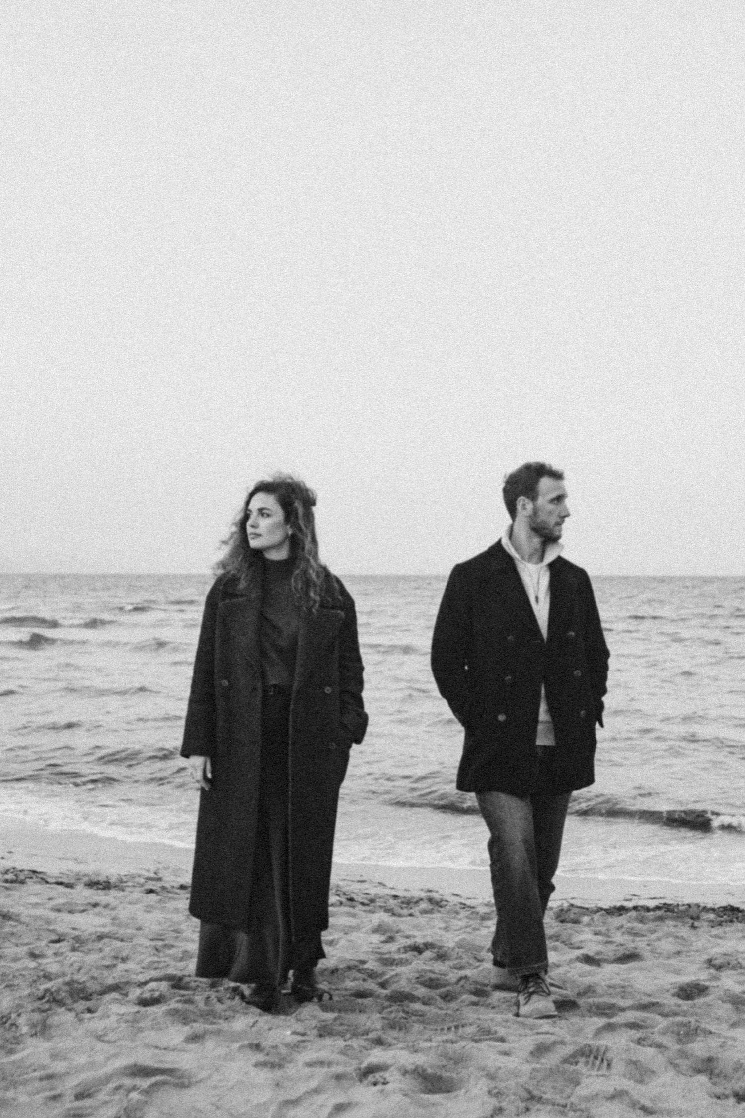 Two people walking along a misty beach shore in black coats, shown in four sequential black and white photographs.