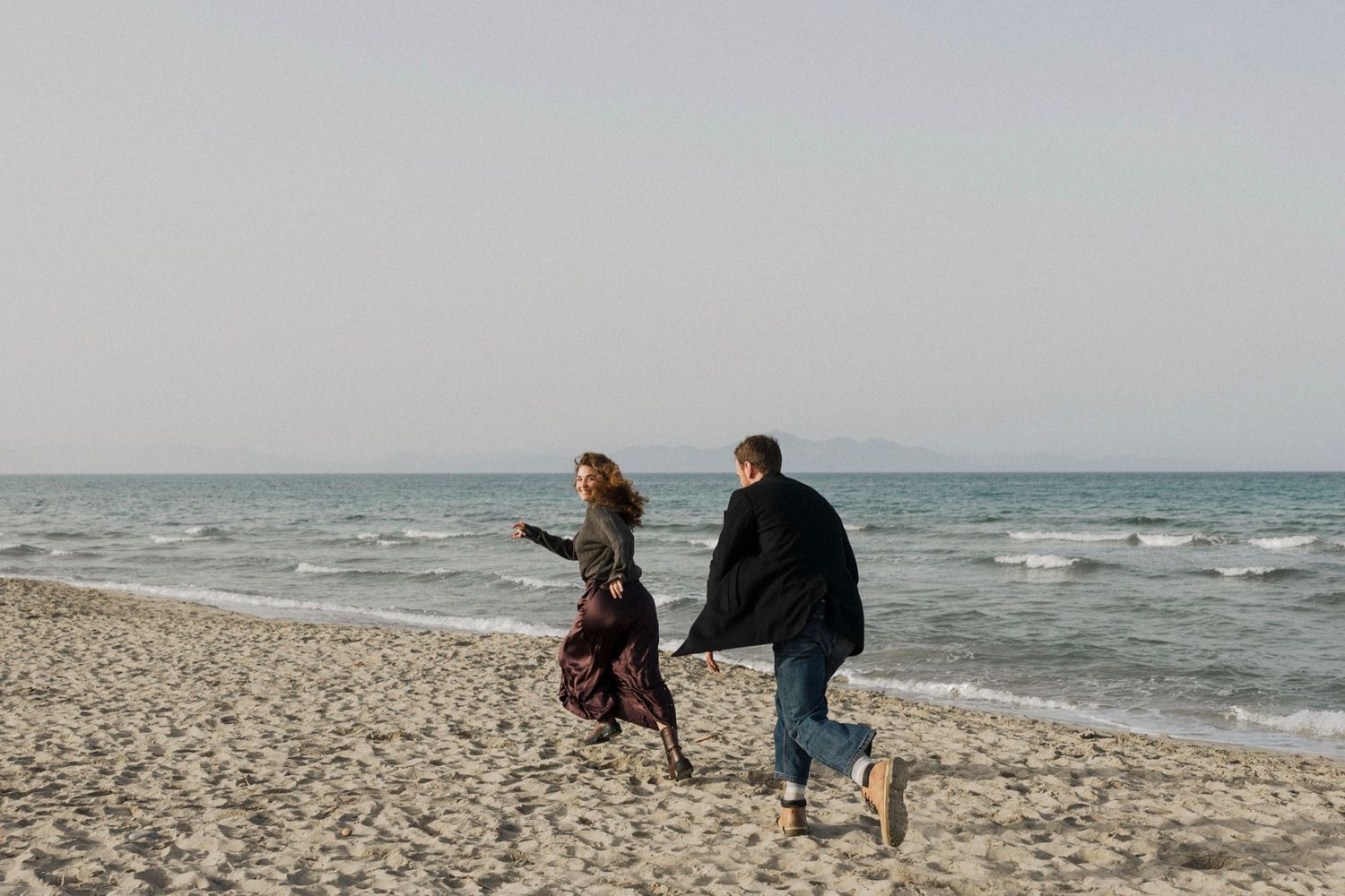 People walking along a sandy beach with rolling waves and cloudy skies in the background.