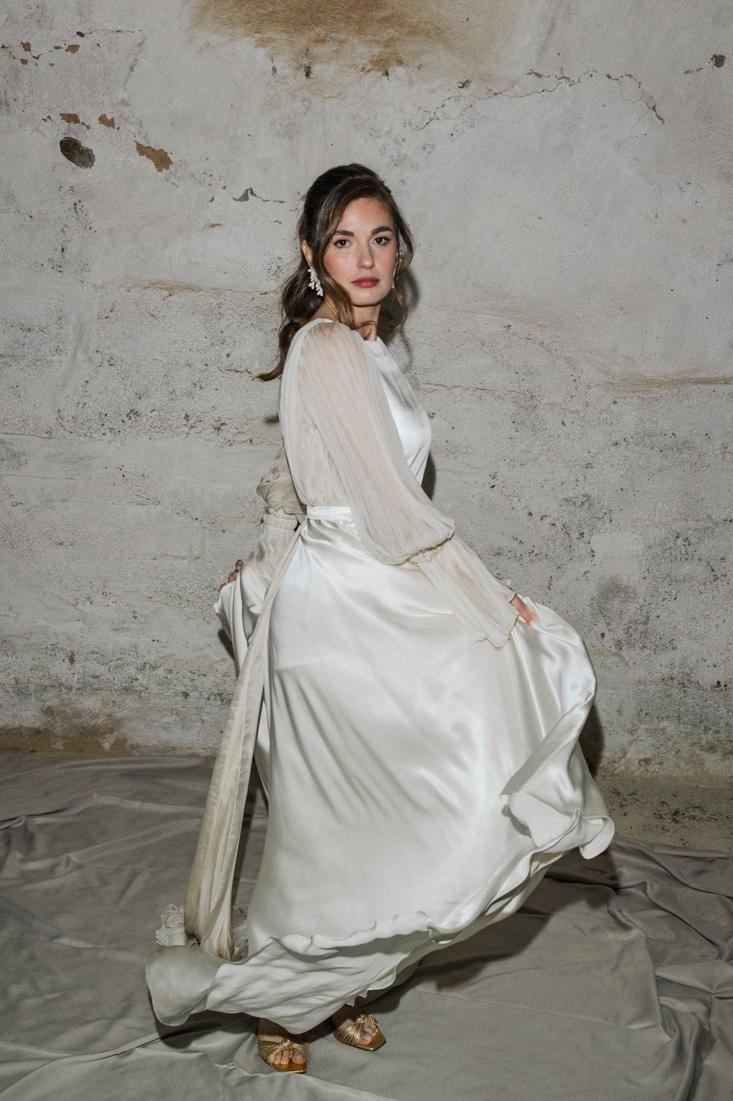 Elegant vintage-inspired white silk wedding dress with long sleeves and flowing skirt photographed against textured wall.