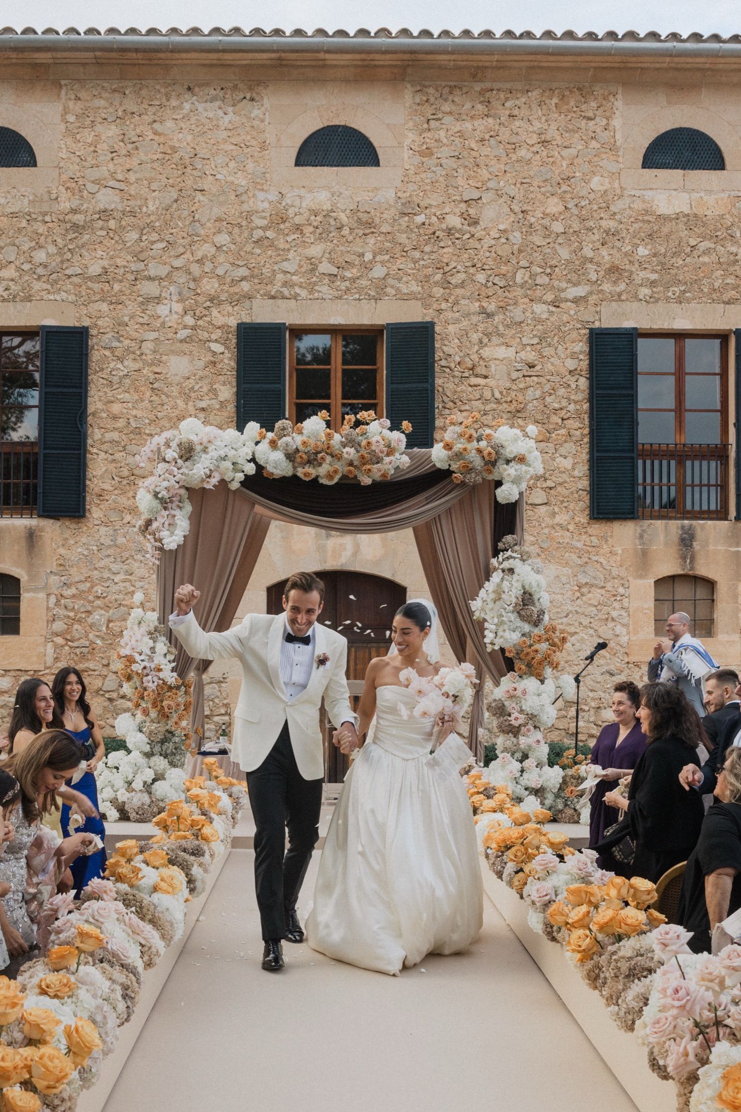 Elegant wedding ceremony setup in a rustic stone courtyard with white and peach floral arrangements and draping decor.