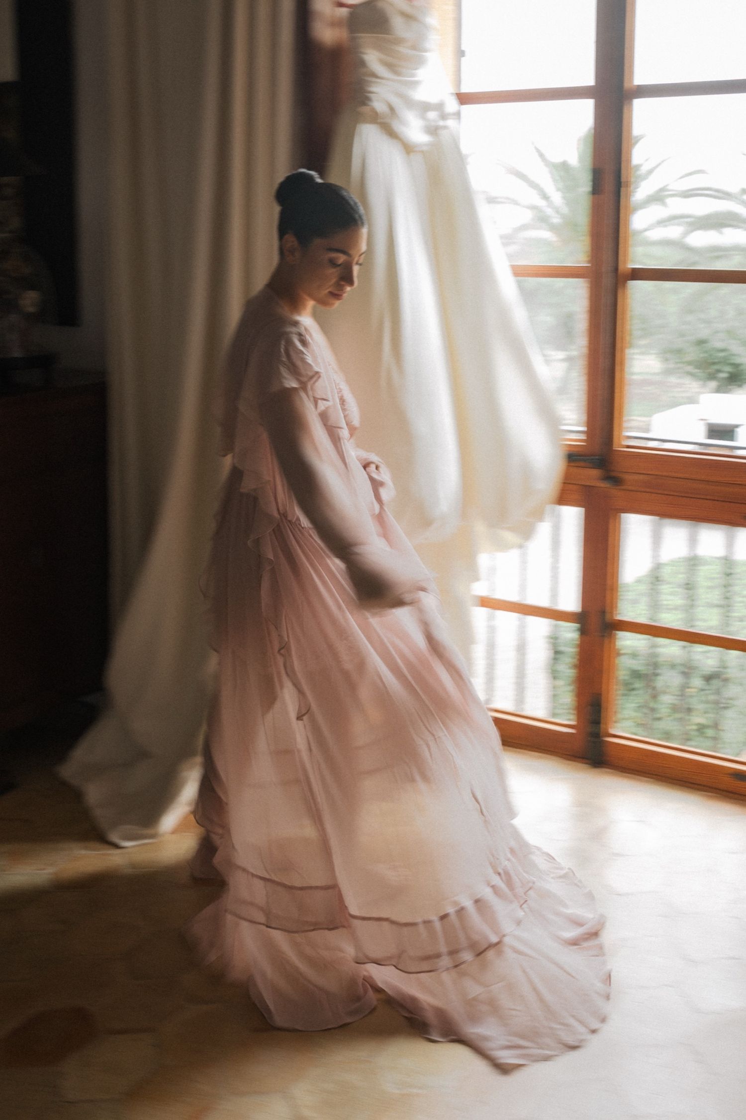 A flowing pink ballgown dress twirls gracefully in front of sunlit windows creating an ethereal bridal moment.