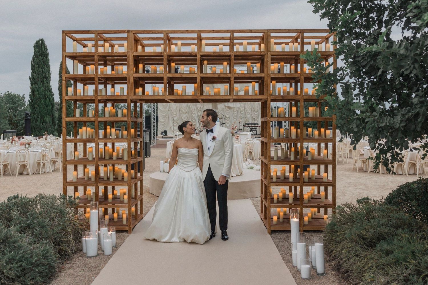 A couple stands under a wooden lattice arch decorated with candles at an outdoor wedding ceremony.