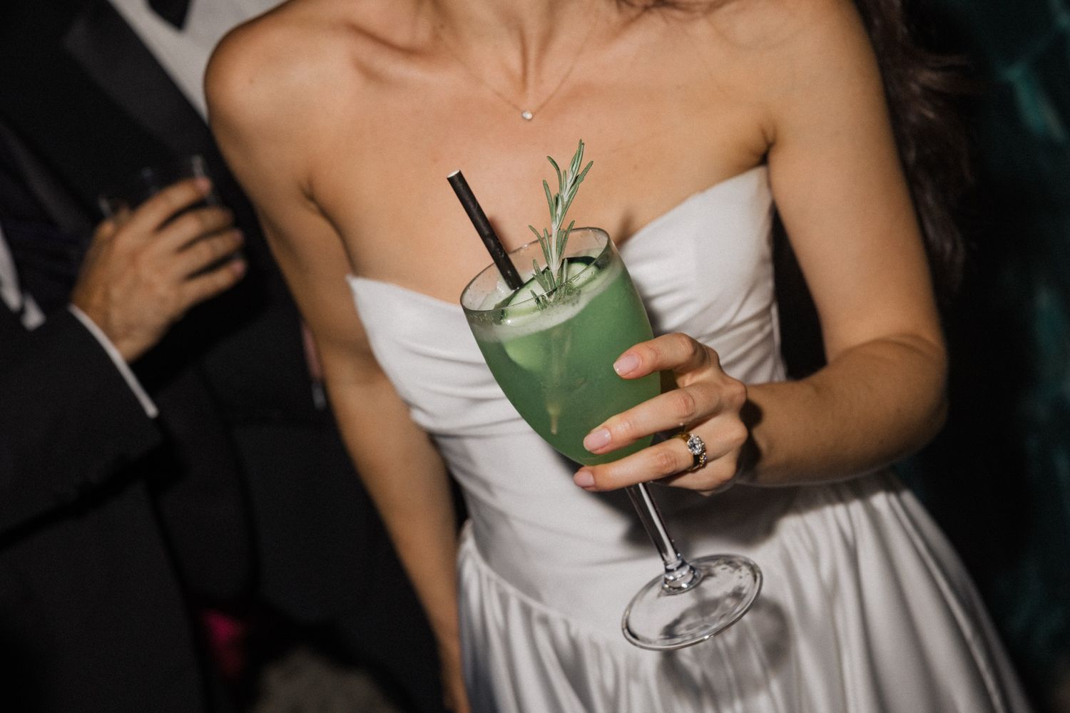 Someone in a white strapless dress holds a green cocktail garnished with herbs at an evening event.
