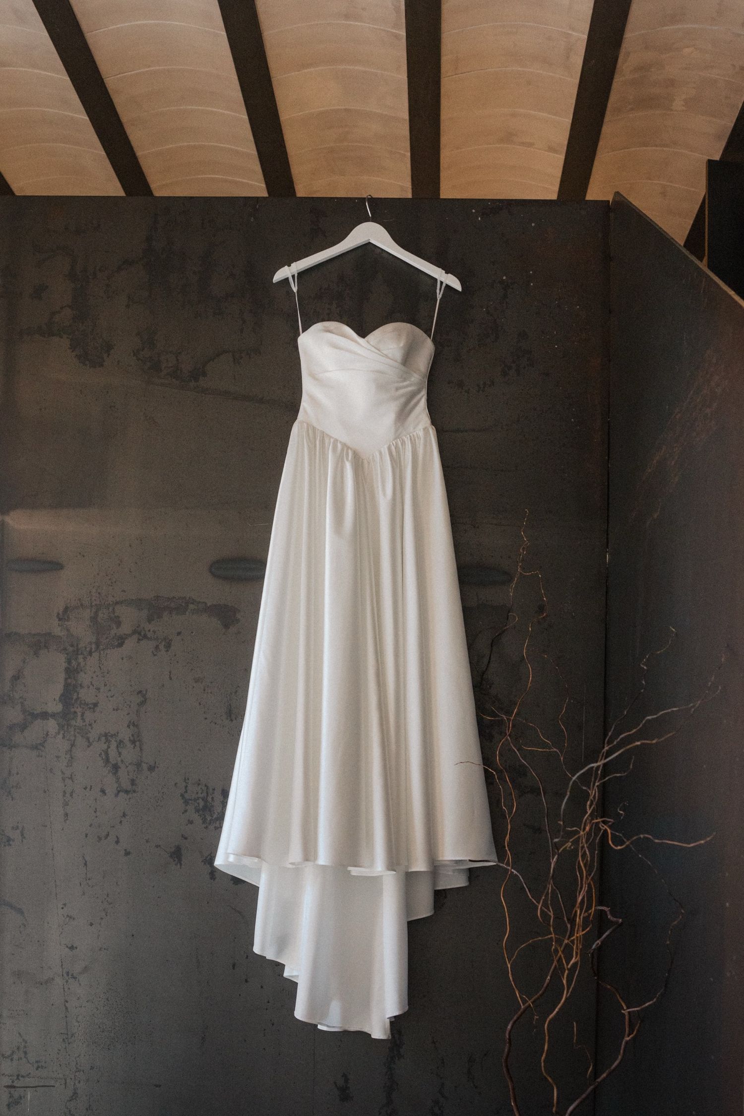 A white flowy dress hangs elegantly against a dark wall with exposed wooden beams visible above.