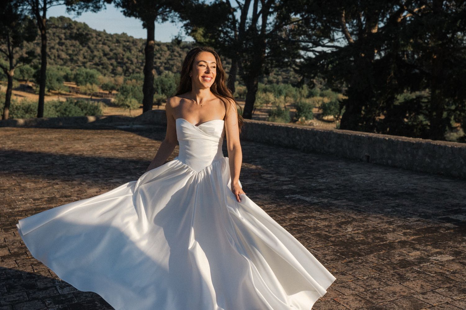 A flowing white wedding dress twirls gracefully in the sunlight against a scenic outdoor backdrop with trees and hills.