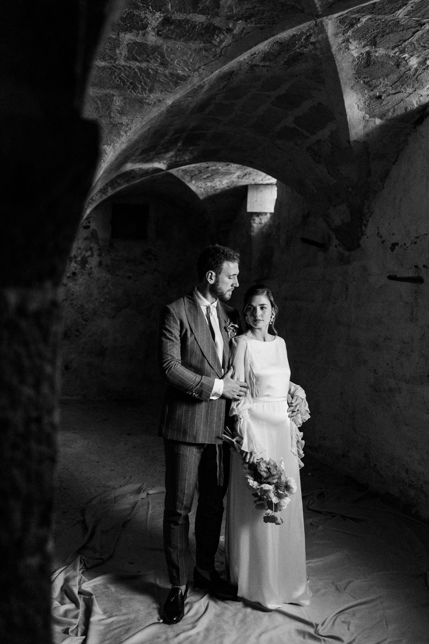 A couple stands together in a dramatic black and white photo under a stone archway.