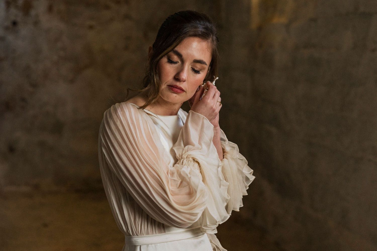 A person in a vintage-inspired white pleated dress with ruffled sleeves poses against a textured brown backdrop.