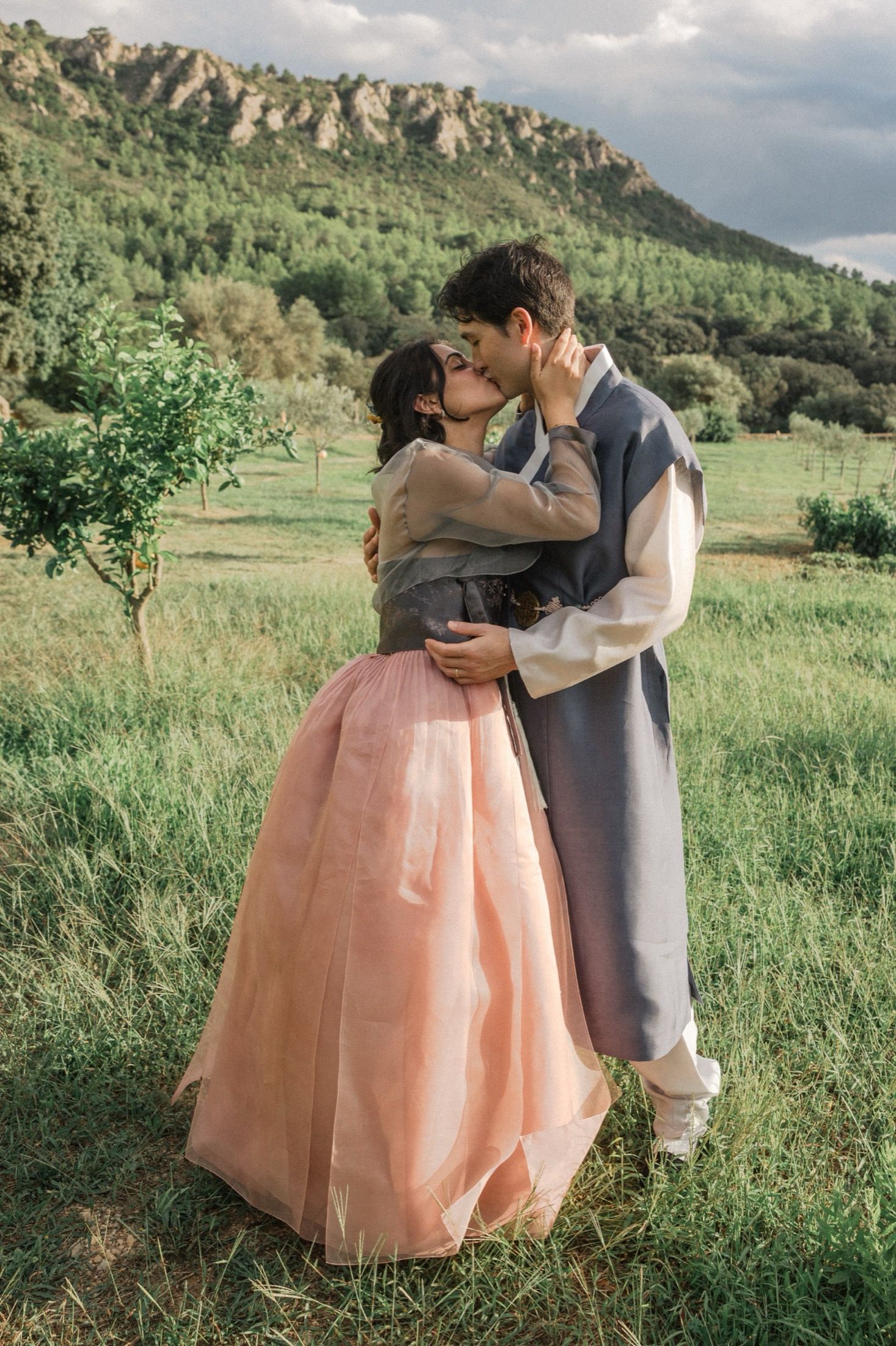 A romantic couple embraces in a meadow with mountains in the background wearing formal attire and a peach-colored gown.