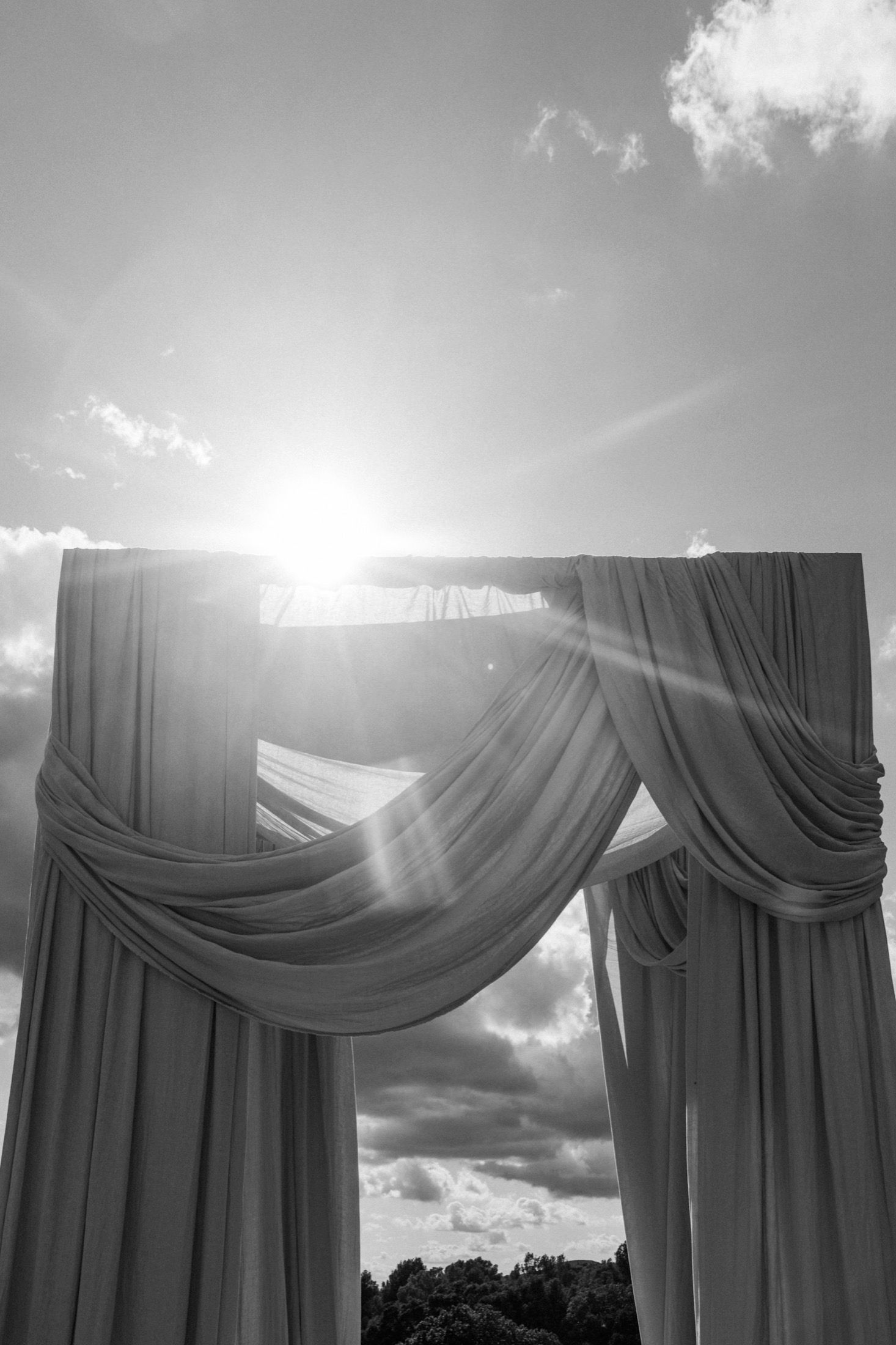 Dramatic black and white photo of draped wedding arch with sunburst shining through fabric against cloudy sky.