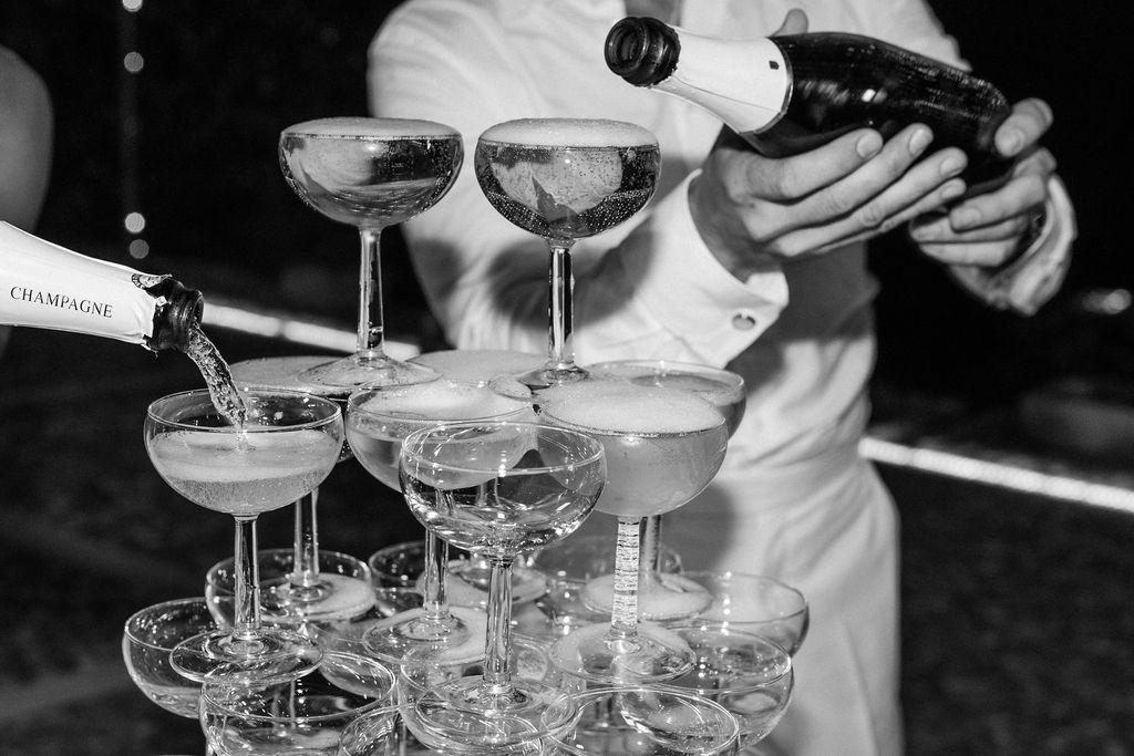 A bartender pours champagne into a tower of stacked coupe glasses creating an elegant cascade effect.