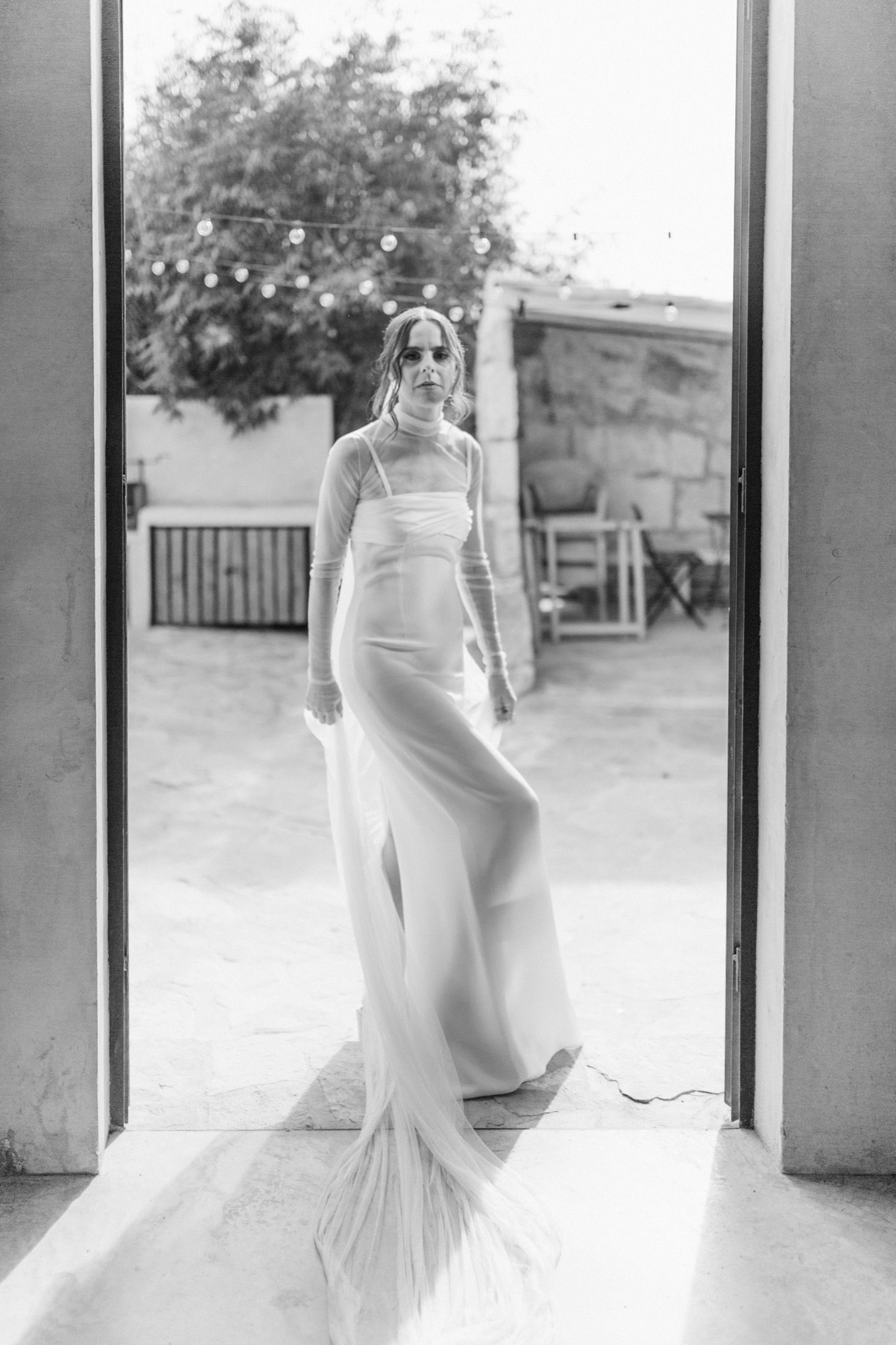 A black and white photograph shows an elegant figure in a flowing white wedding gown standing in a doorway.
