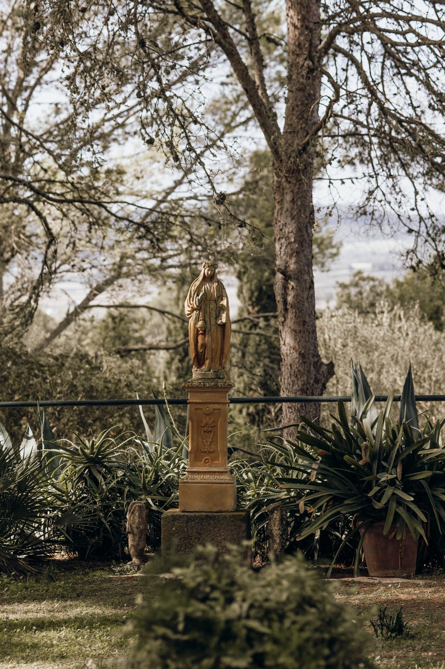 Rustic wooden sculptures and art pieces displayed among tropical plants and trees in an outdoor garden setting.