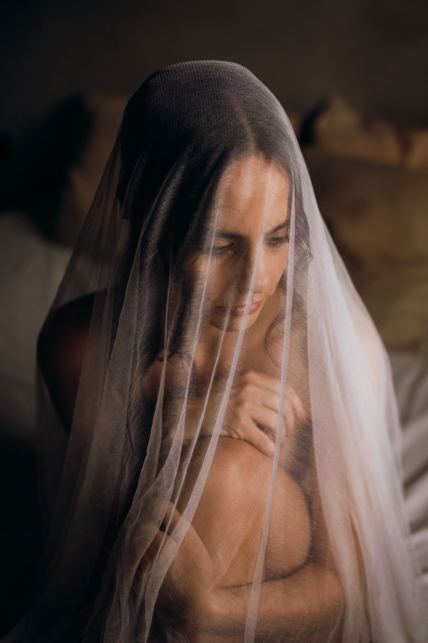 A bride wearing a delicate white veil looks down with a serene expression in a moody, artistic portrait series.