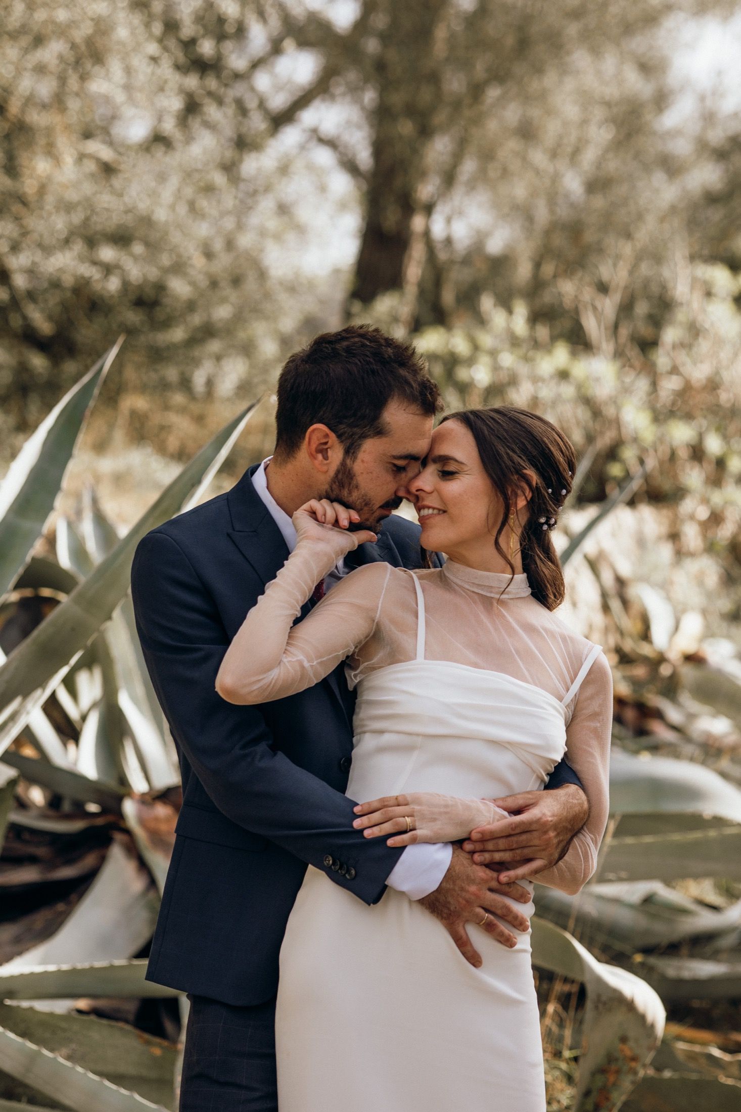 A romantic couple embraces outdoors in a garden setting with soft lighting and agave plants in the background.