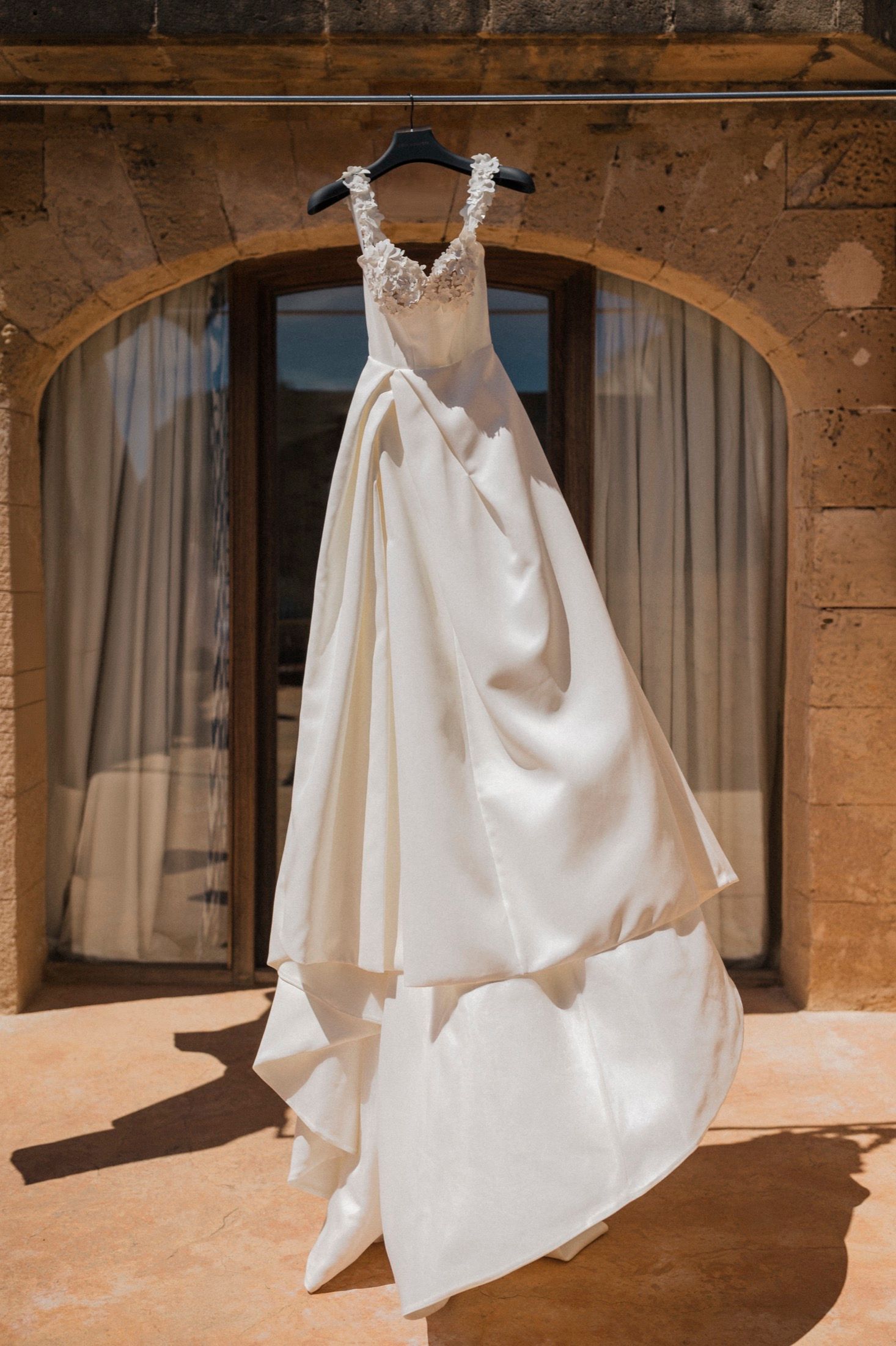 Elegant white wedding dress with lace bodice hanging on a hanger against a stone archway window.