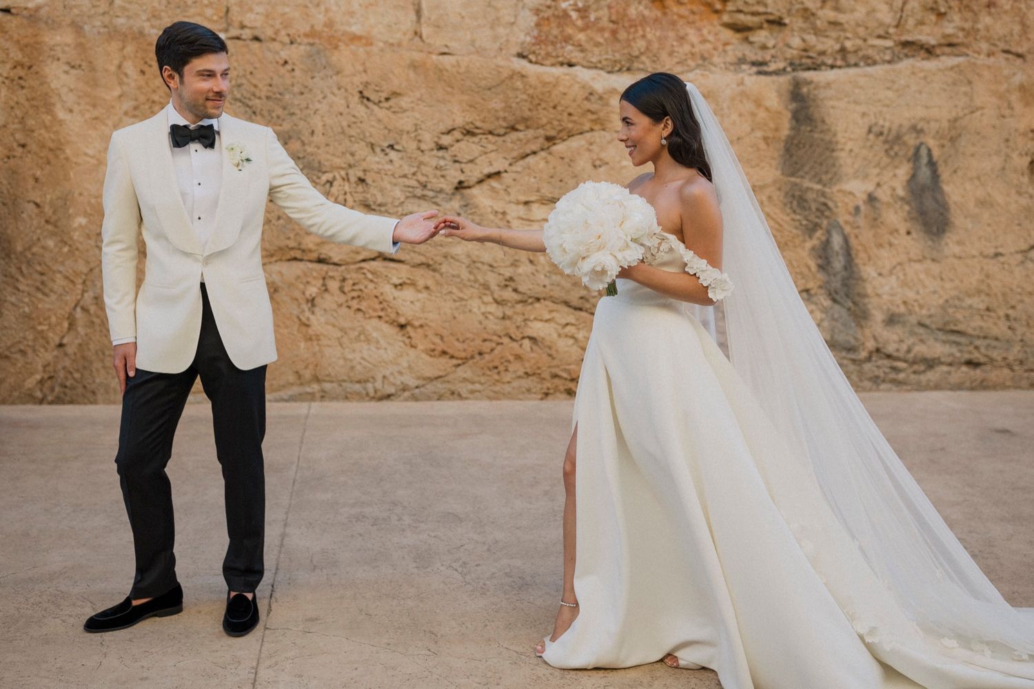 Bride in white gown holding bouquet and groom in white tuxedo jacket hold hands at outdoor wedding venue.