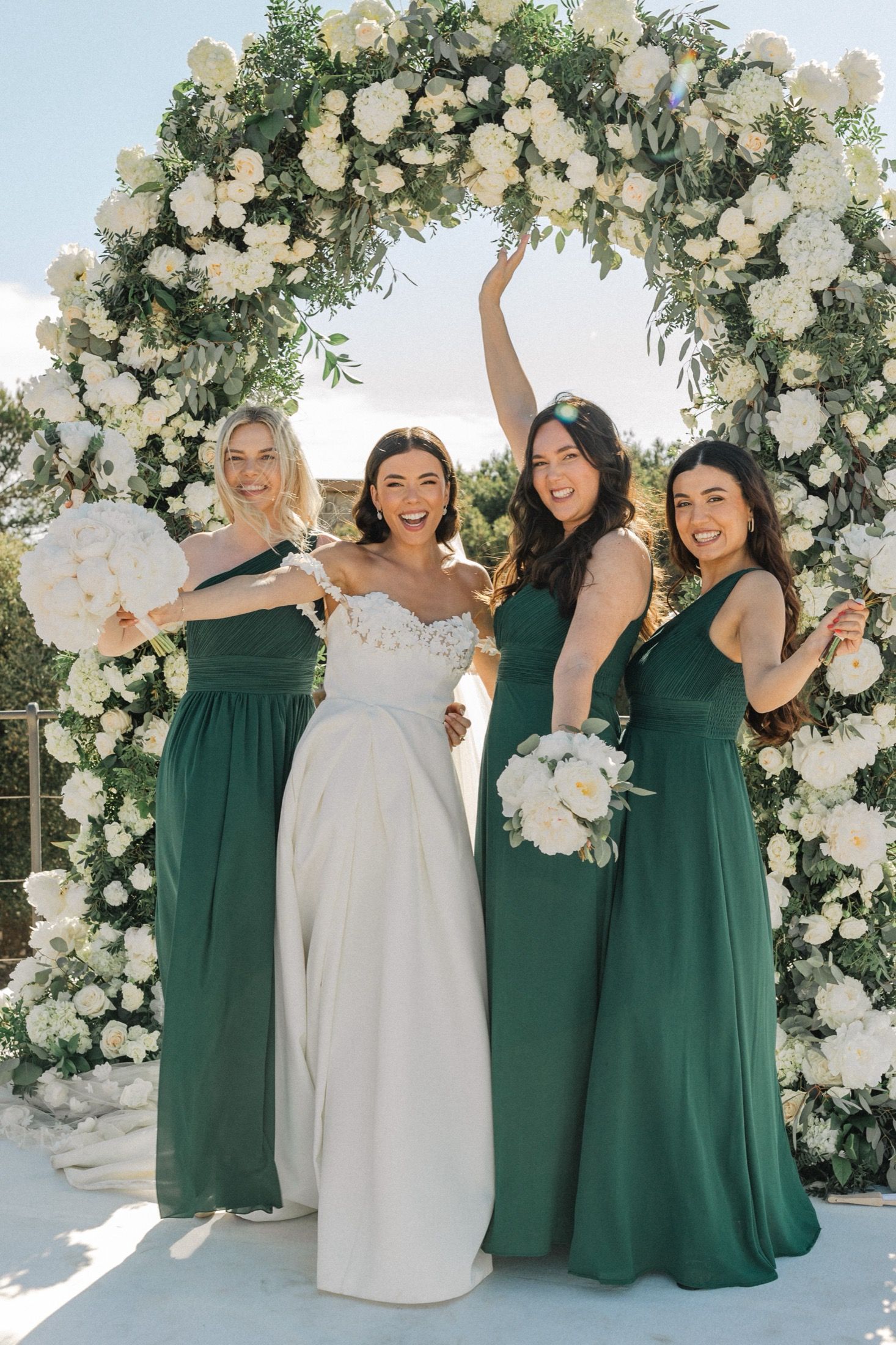 Bride in white gown with three bridesmaids in emerald green dresses under floral arch with white roses.