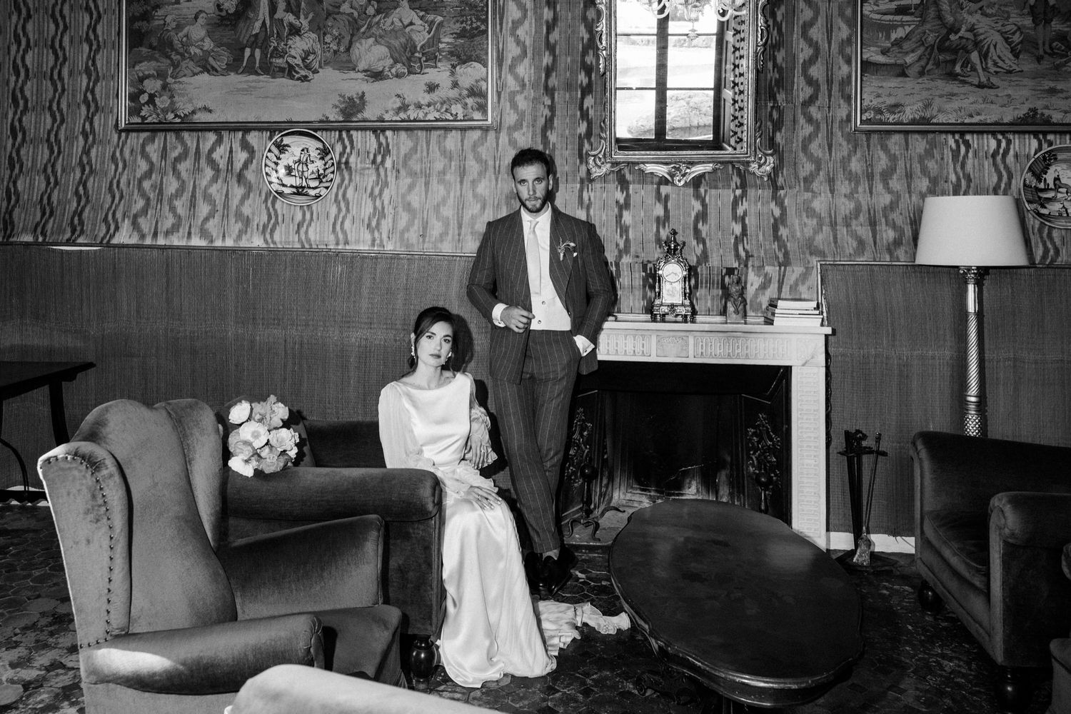 Elegant black and white photo of a couple in formal attire posing in a vintage-style living room with ornate wallpaper.