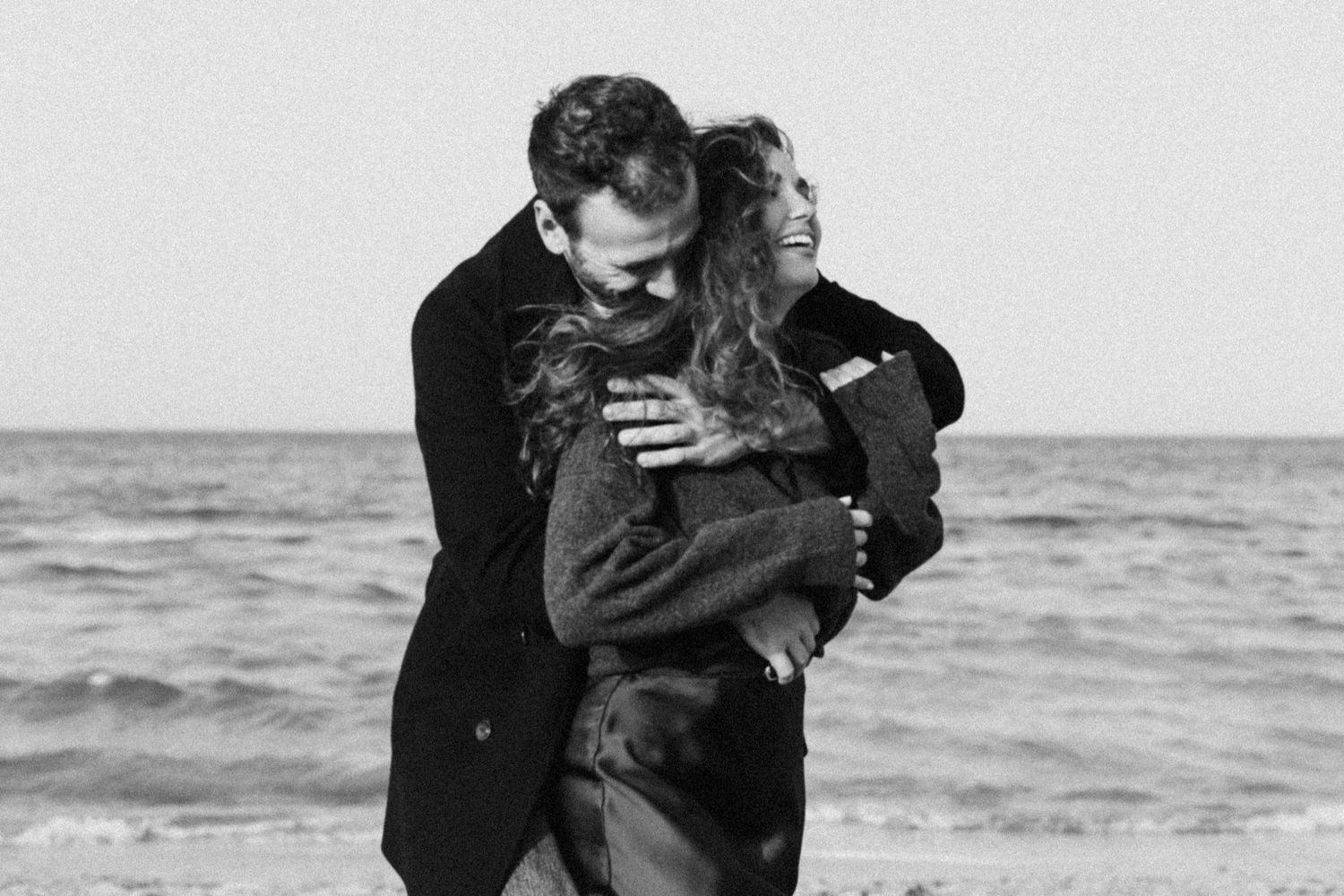 A romantic couple embraces on the beach in black and white as waves crash behind them.