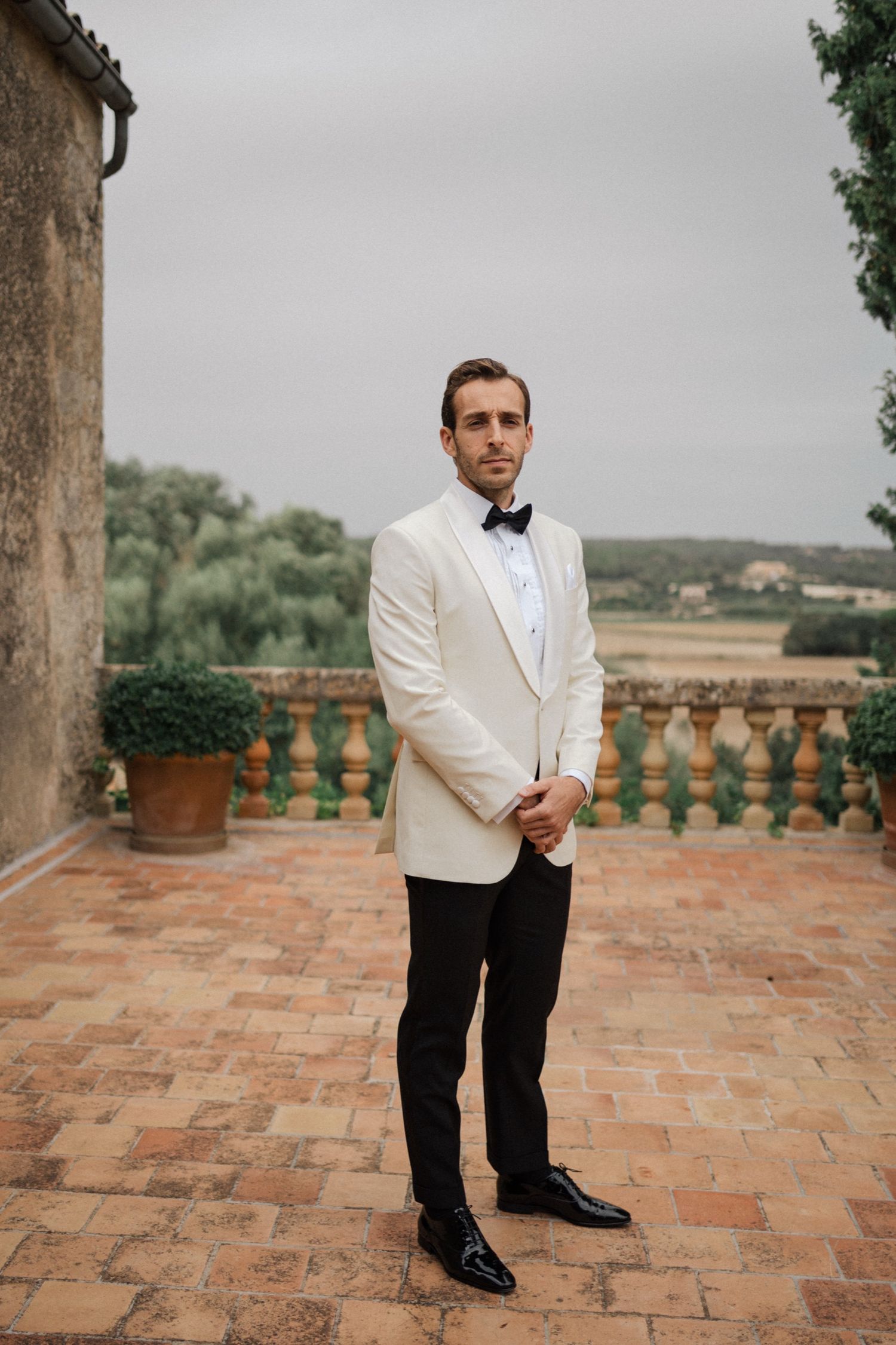 A groom in a white dinner jacket and bow tie stands on a rustic terrace overlooking countryside in Spain.