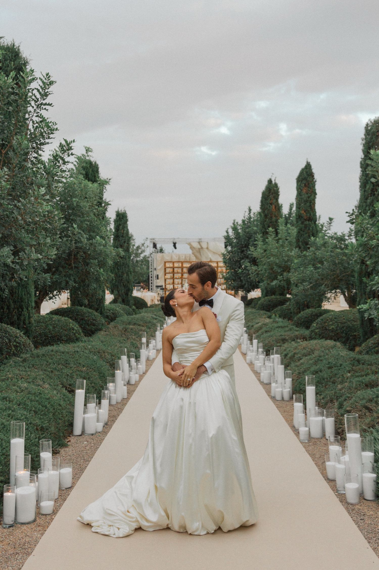 A romantic outdoor wedding ceremony setup with a white aisle runner lined with glowing candles and cypress trees.