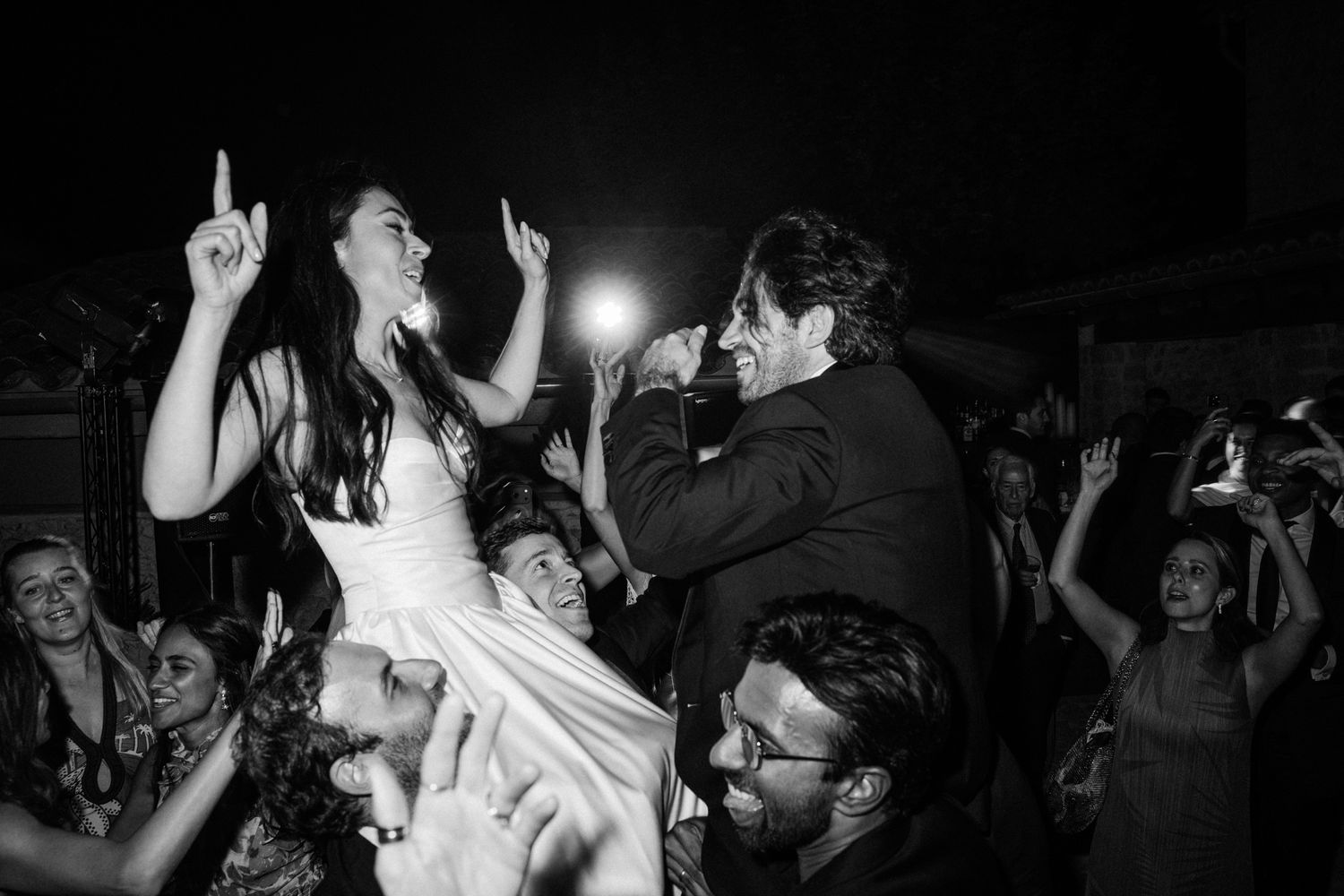 Silhouetted crowd of people dancing with raised arms at a nightclub party with bright lights in background.