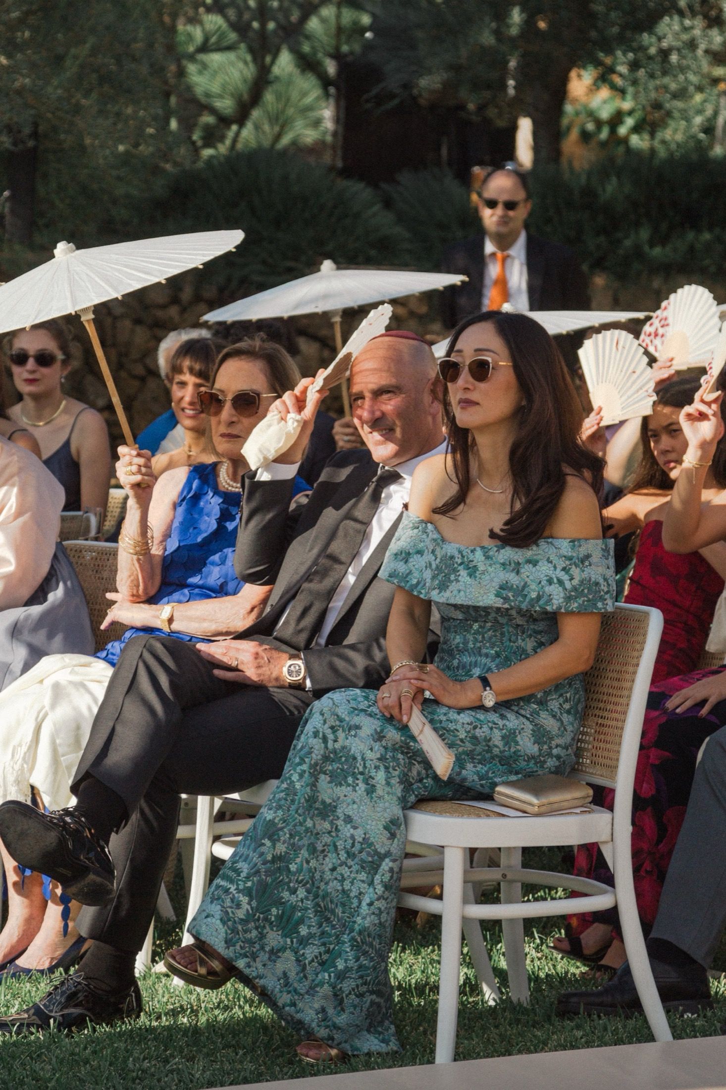 Wedding guests hold white parasols to shade themselves while sitting in white chairs during an outdoor ceremony.