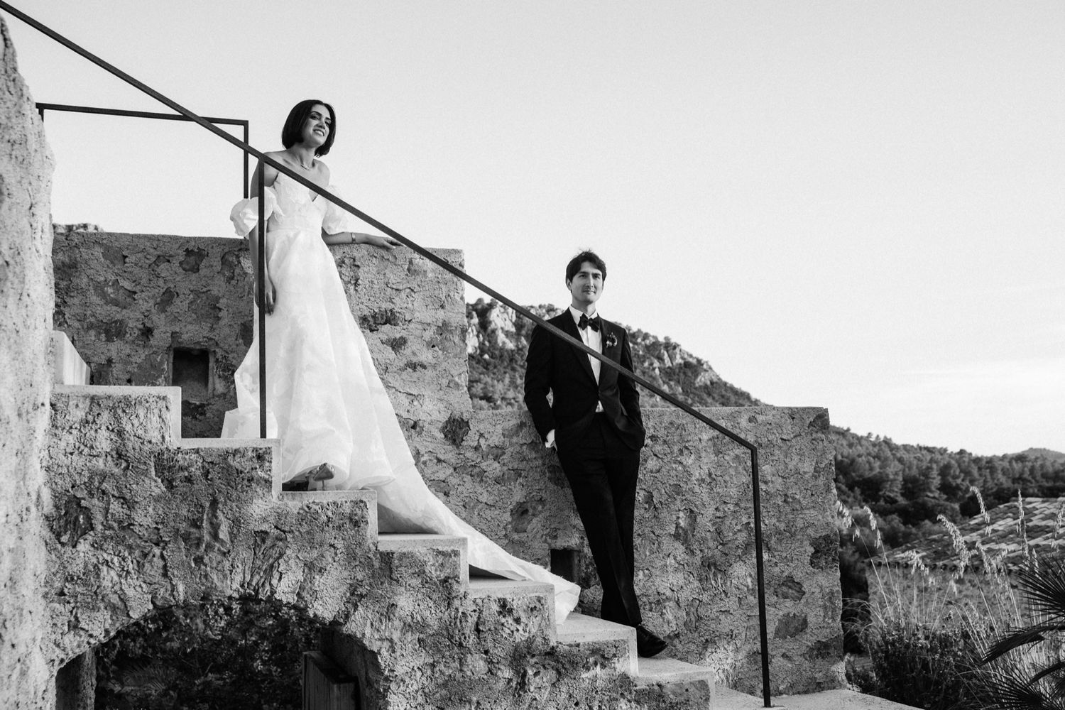 A bride in a flowing white gown and groom stand on rustic stone steps for wedding portraits.