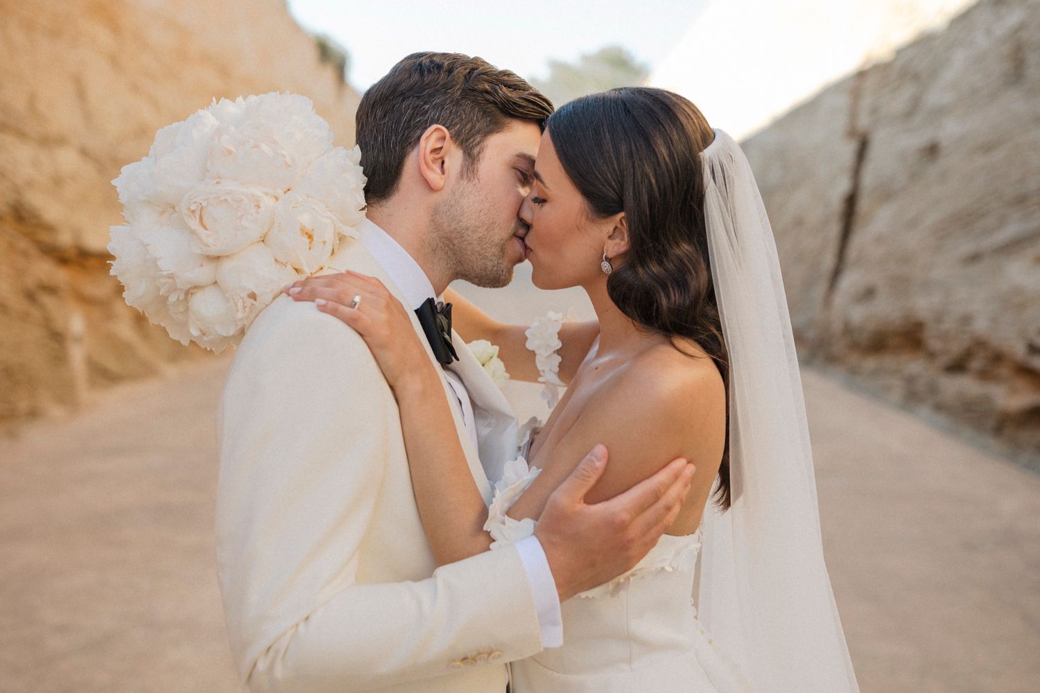 Bride and groom share a romantic kiss outdoors, bride holding white floral bouquet, both dressed in elegant wedding attire.