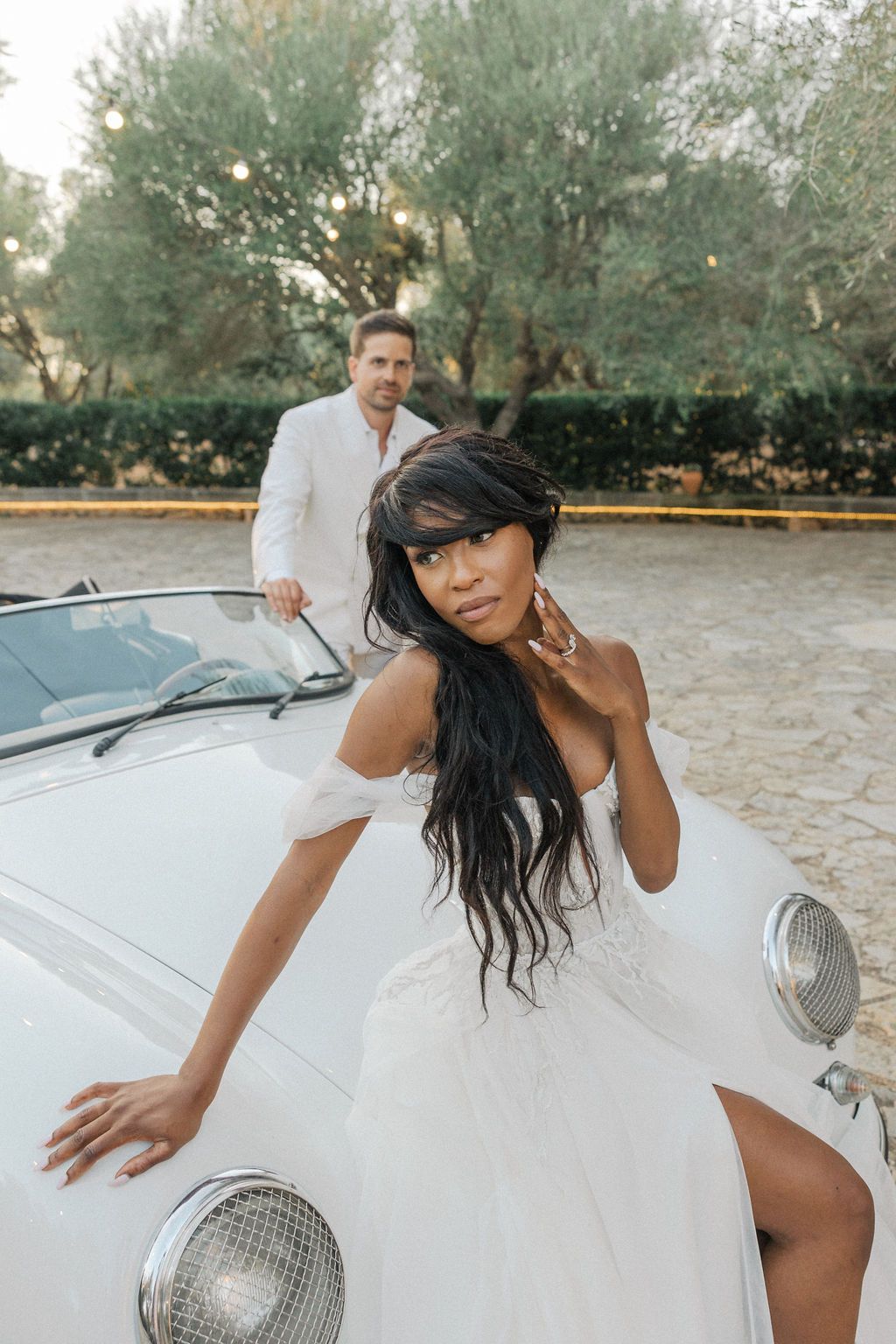 Elegant couple in white attire posing with classic car during outdoor photoshoot.