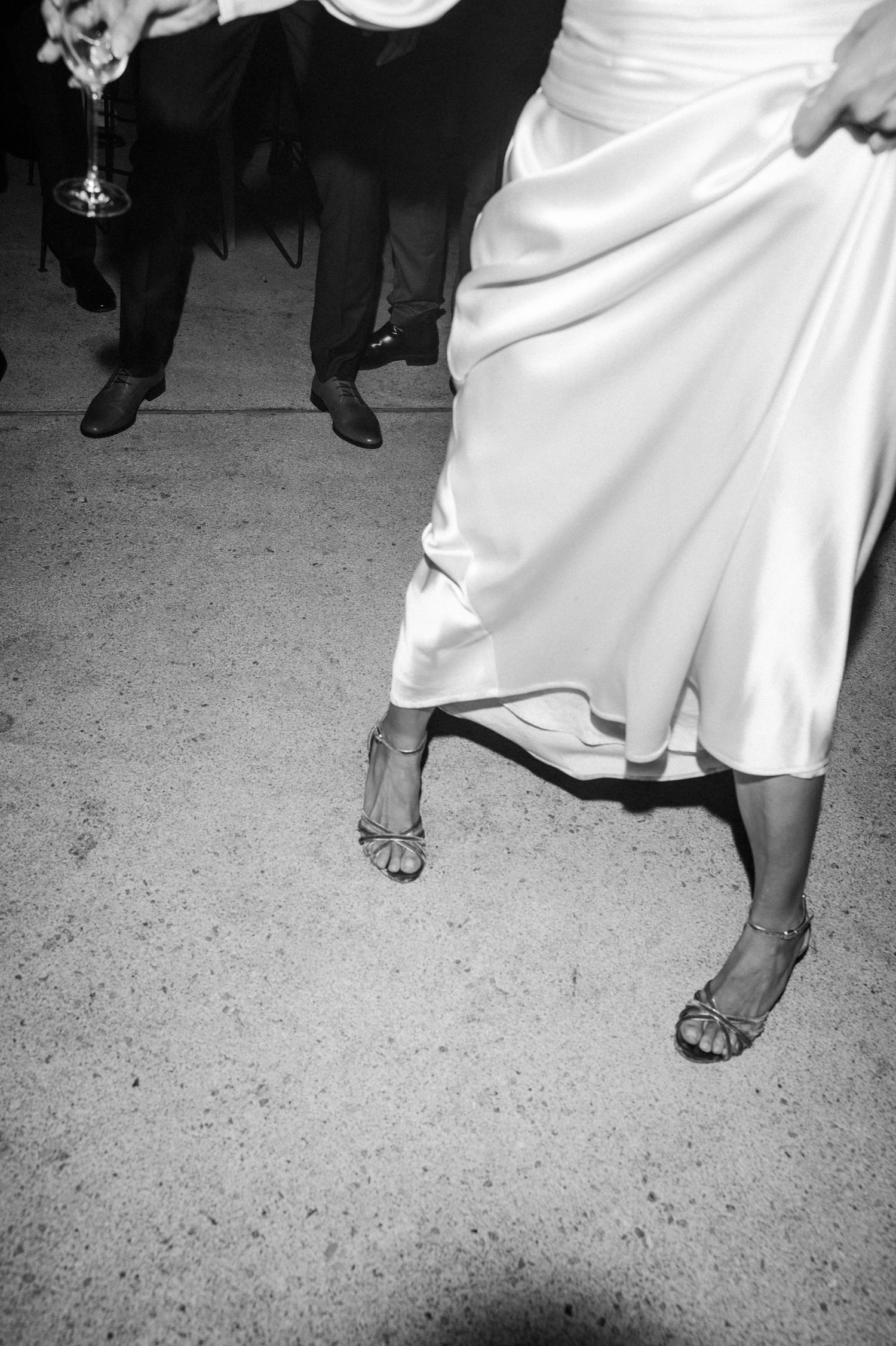 Black and white photo showing dancing feet and flowing white dress on a concrete floor.