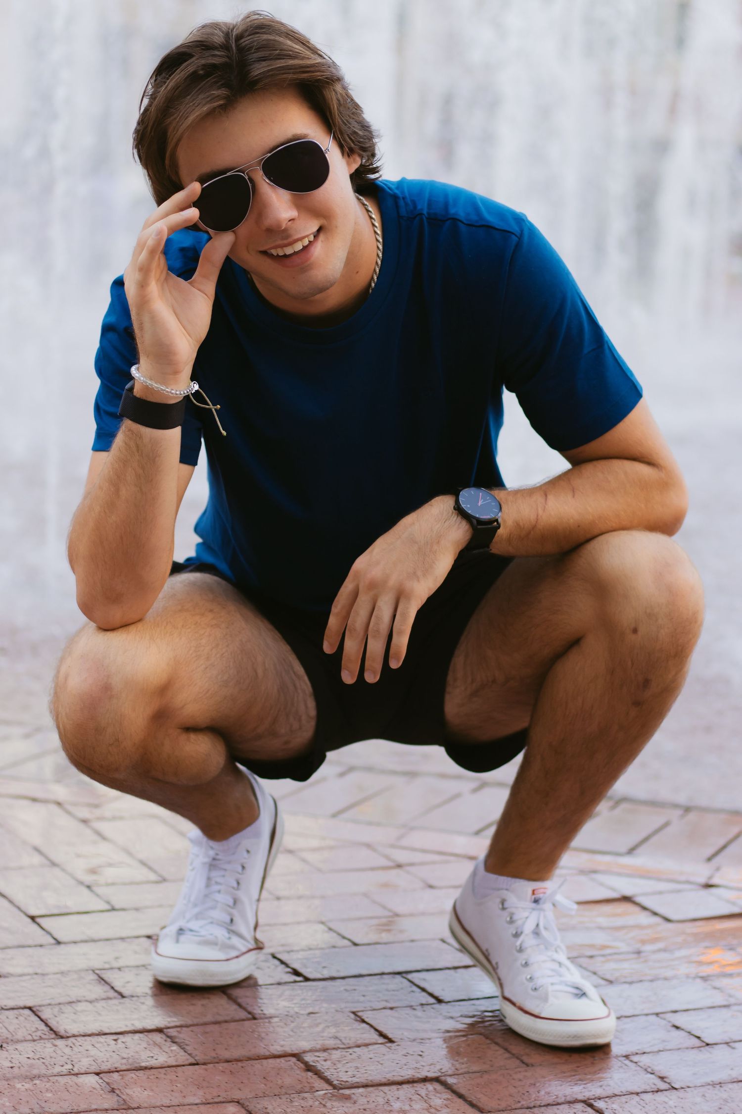Young person in blue t-shirt and shorts squatting while wearing sunglasses and white sneakers.