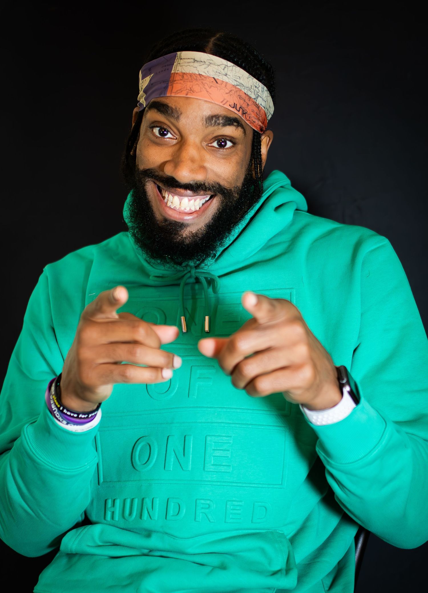 Portrait of smiling person wearing green hoodie and patterned headband against black background.